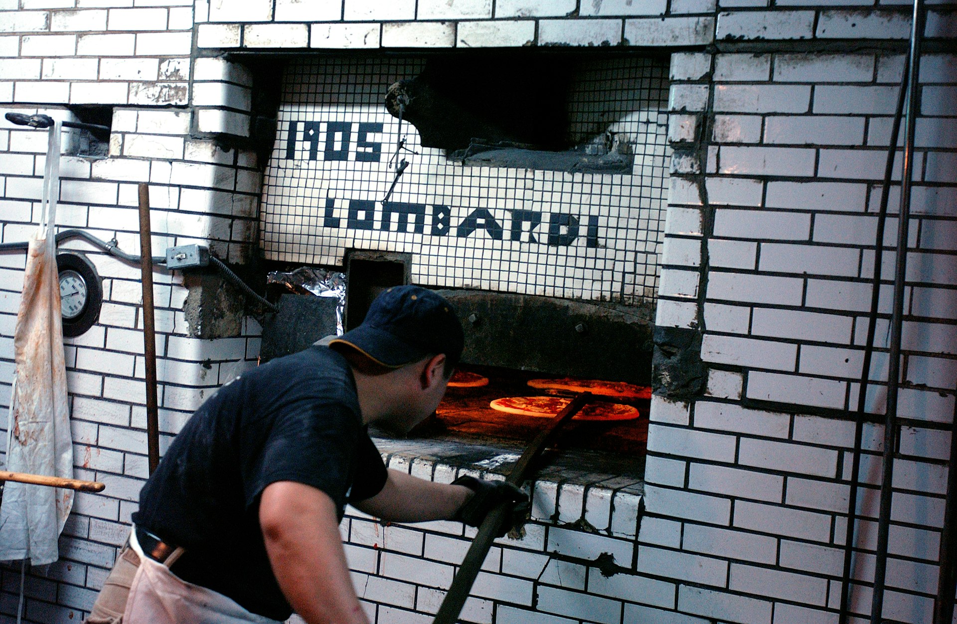 A man pushes an uncooked pizza pie into a subway tiled oven. Above are the words '1905 Lombardi' 