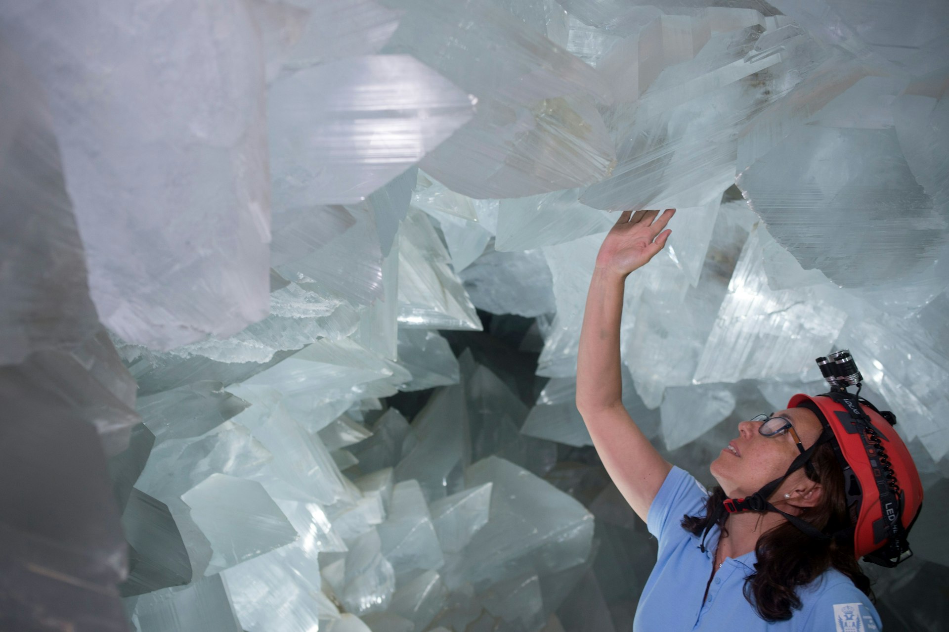 A woman wearing a red helmet with a torchlight attached touches a large white crystal descending from a ceiling. She's surrounded by large crystals inside the la geoda de pulpi