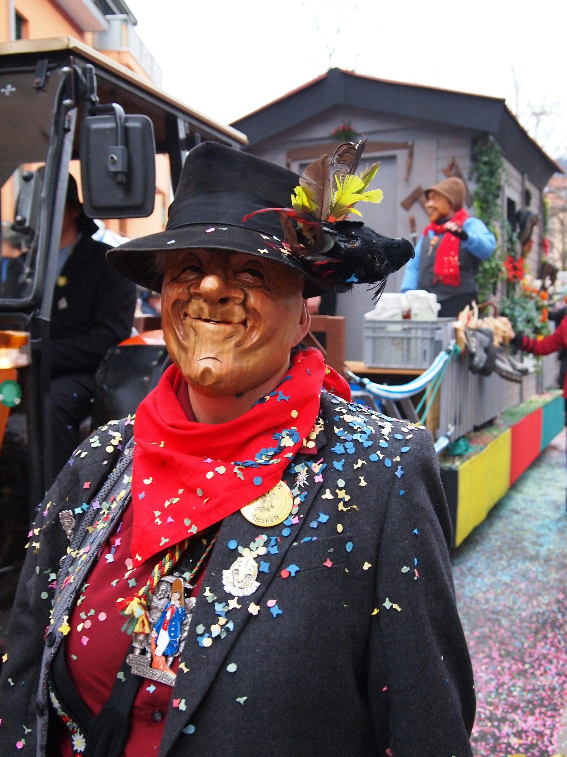A person dressed formally covered in confetti wears a smiling wooden face mask