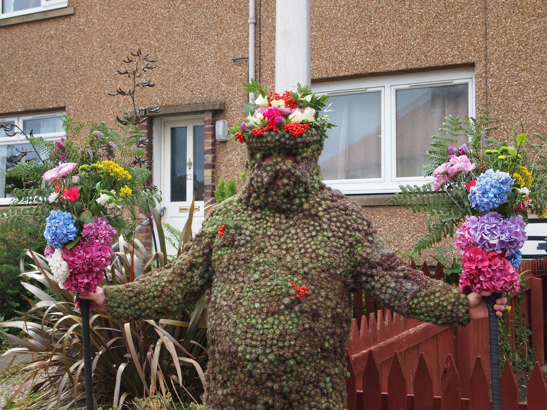 A person completely covered in a costume of dark green seeds, holding bunches of flowers