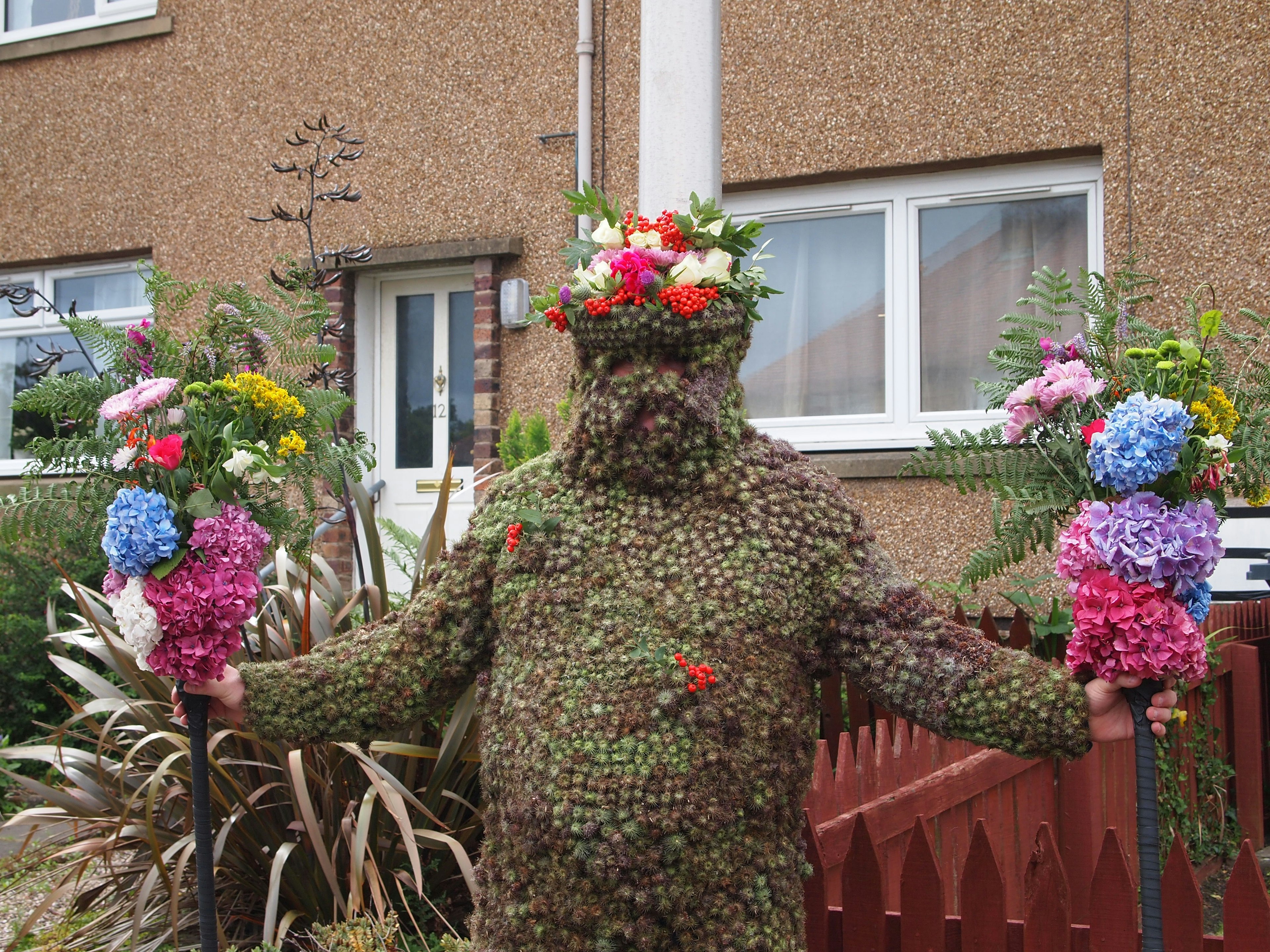 A person completely covered in a costume of dark green seeds, holding bunches of flowers