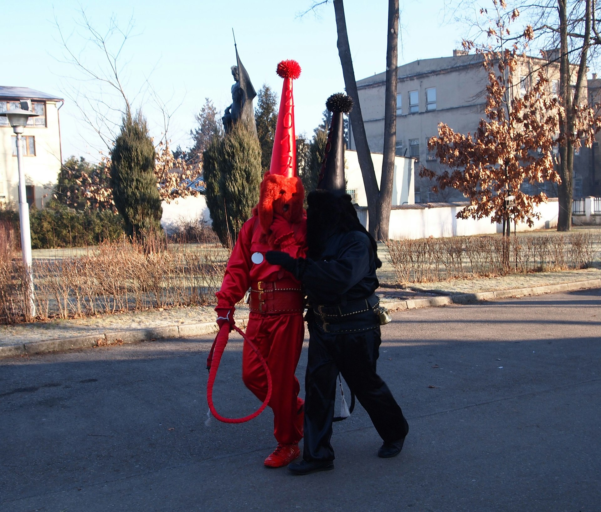One person dressed head-to-toe in red, and another in black, wear pointy hats and hold whips