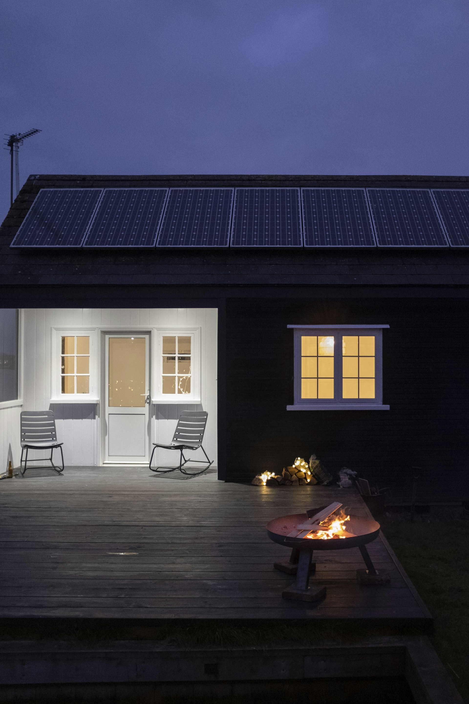 An exterior shot of a cabin's deck at dusk, with two chairs and a fire burning in a fire pit