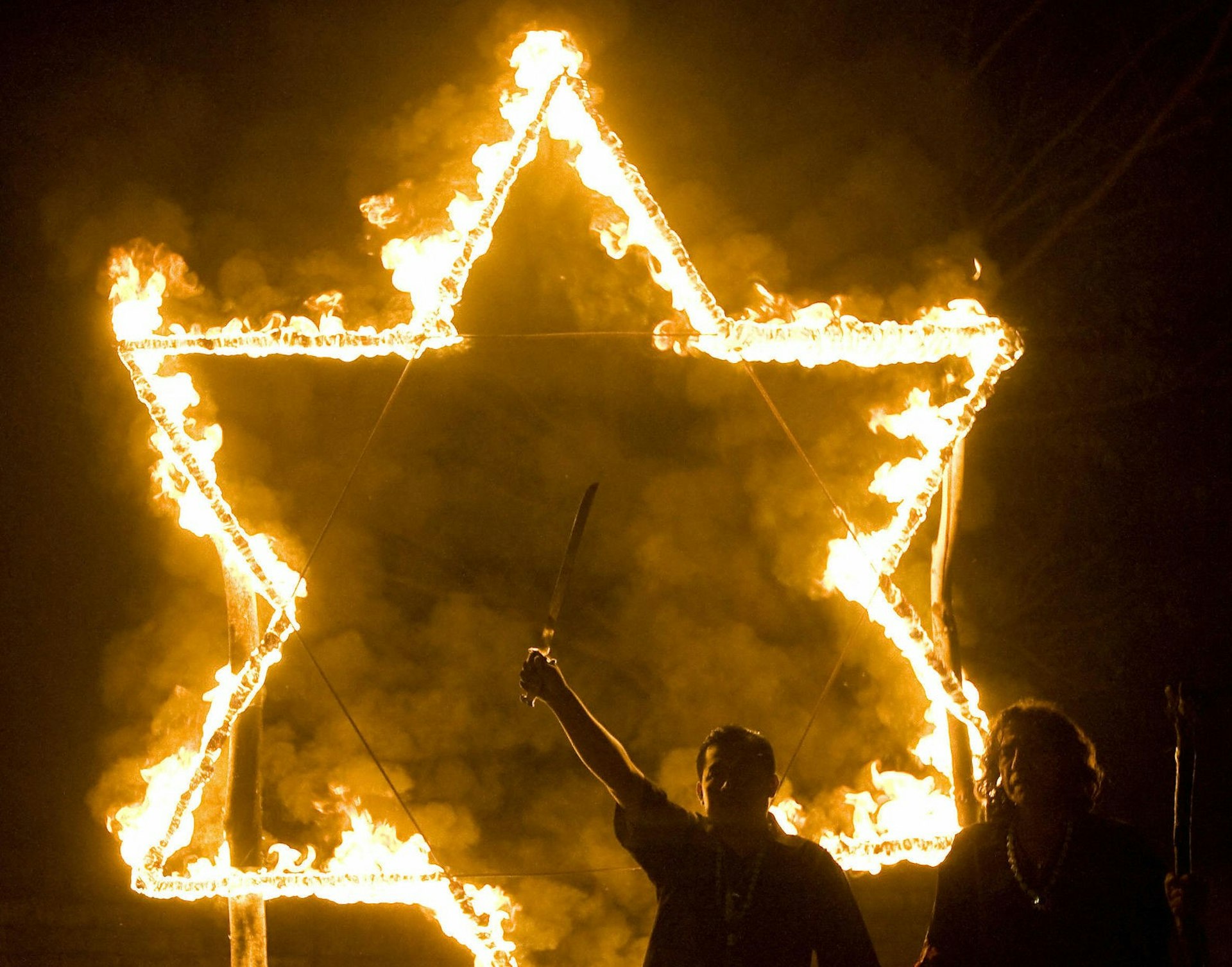 A burning pentagram with two men standing in front of it in the dark, one is raising a sword.
