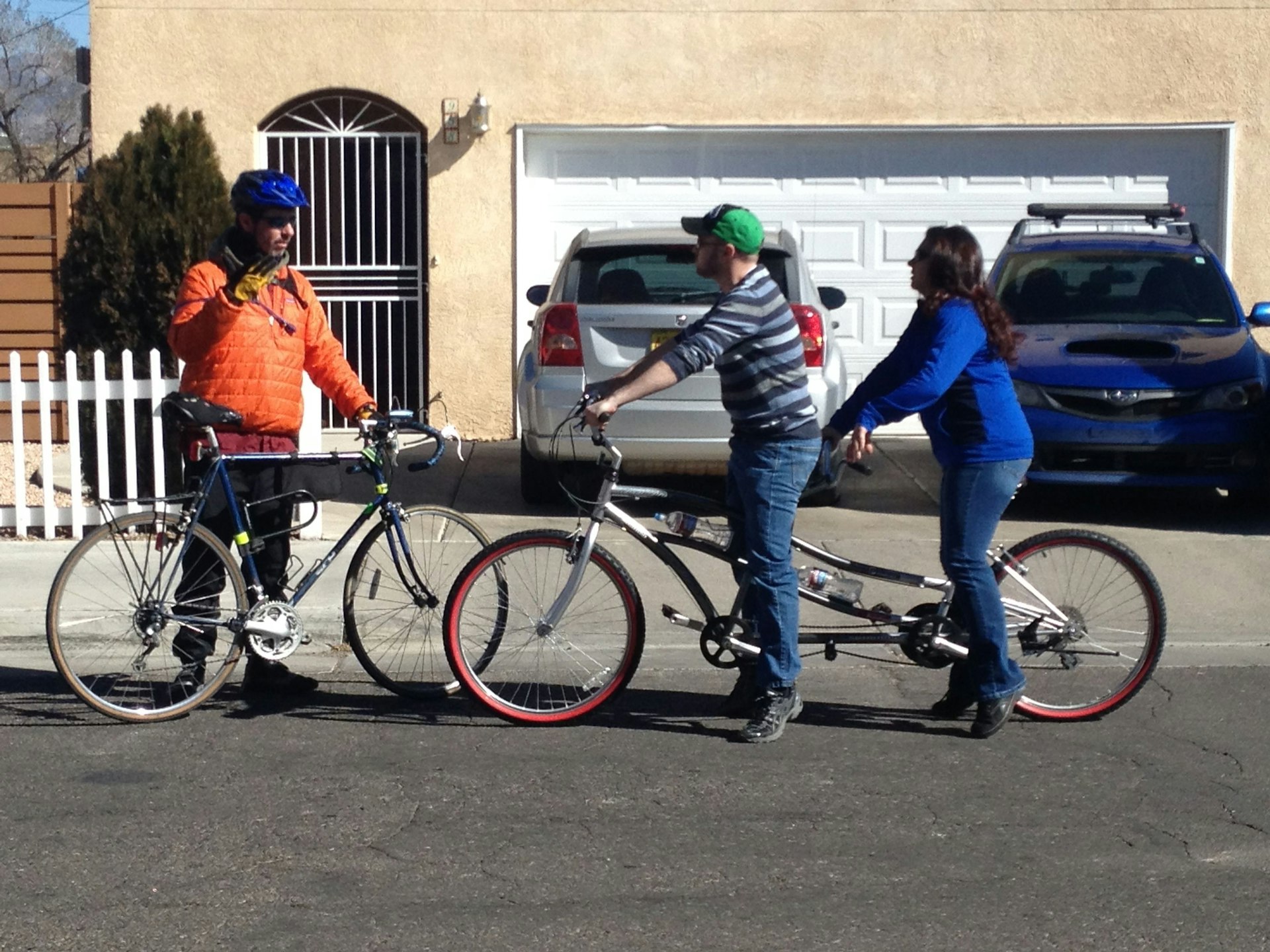 A man with a bicycle on the left side of the photo speaks with and gestures to two adults on a tandem bicycle that runs perpendicular to the viewer across the rest of the frame