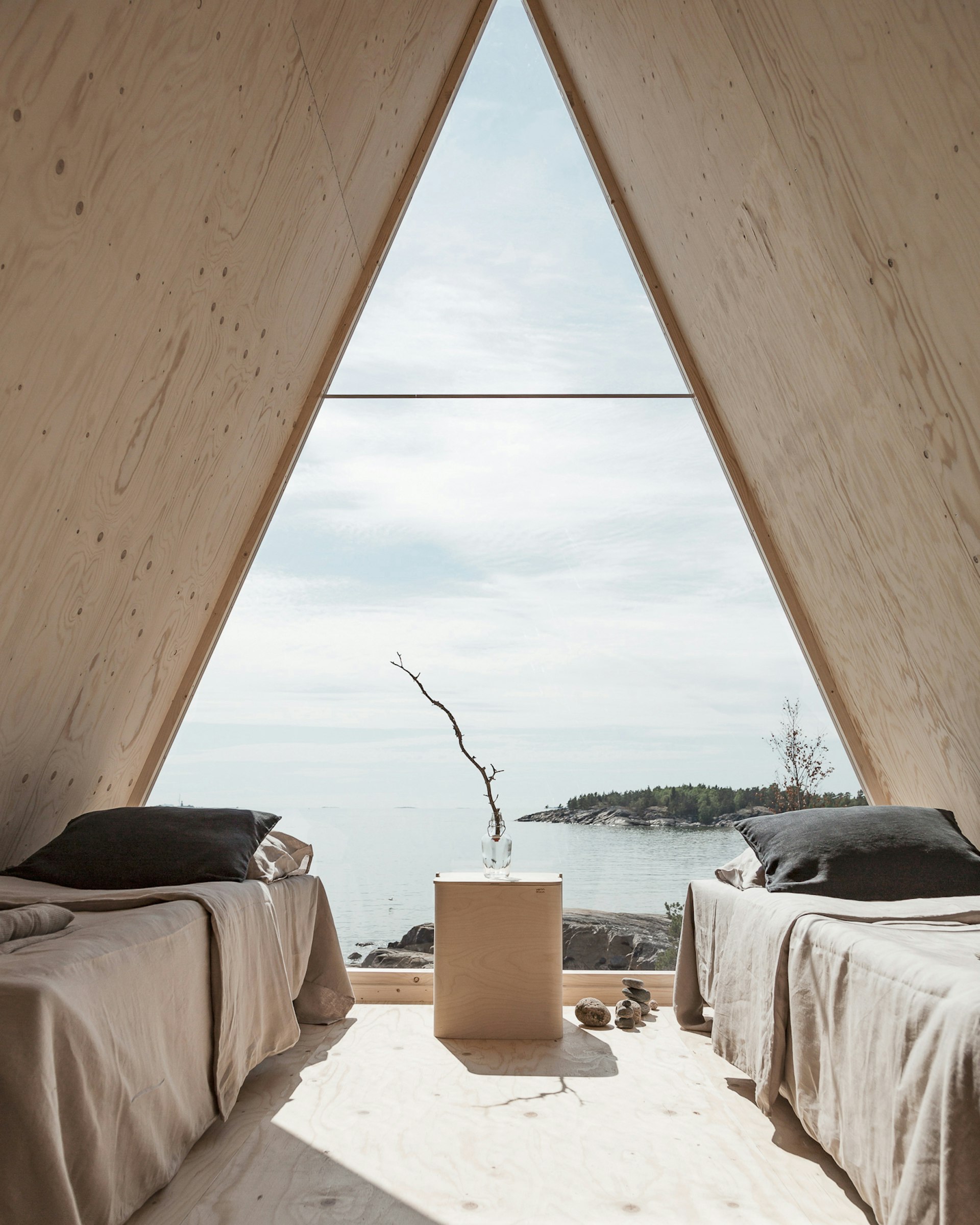 A view to the sea framed by the door of a triangular cabin, with a single bed on each side