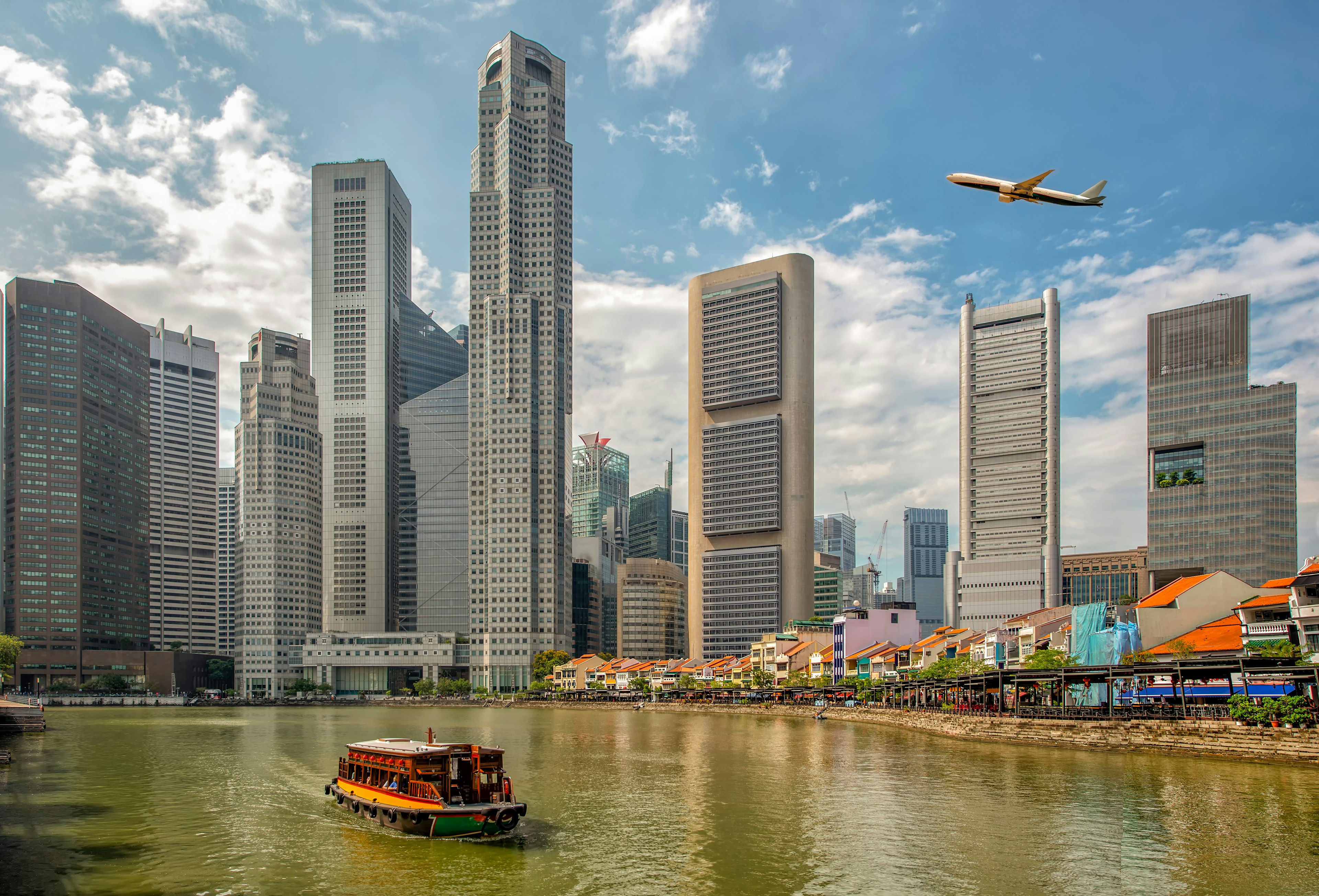 A plane flies over downtown Singapore's skyline with a ferry on the river below