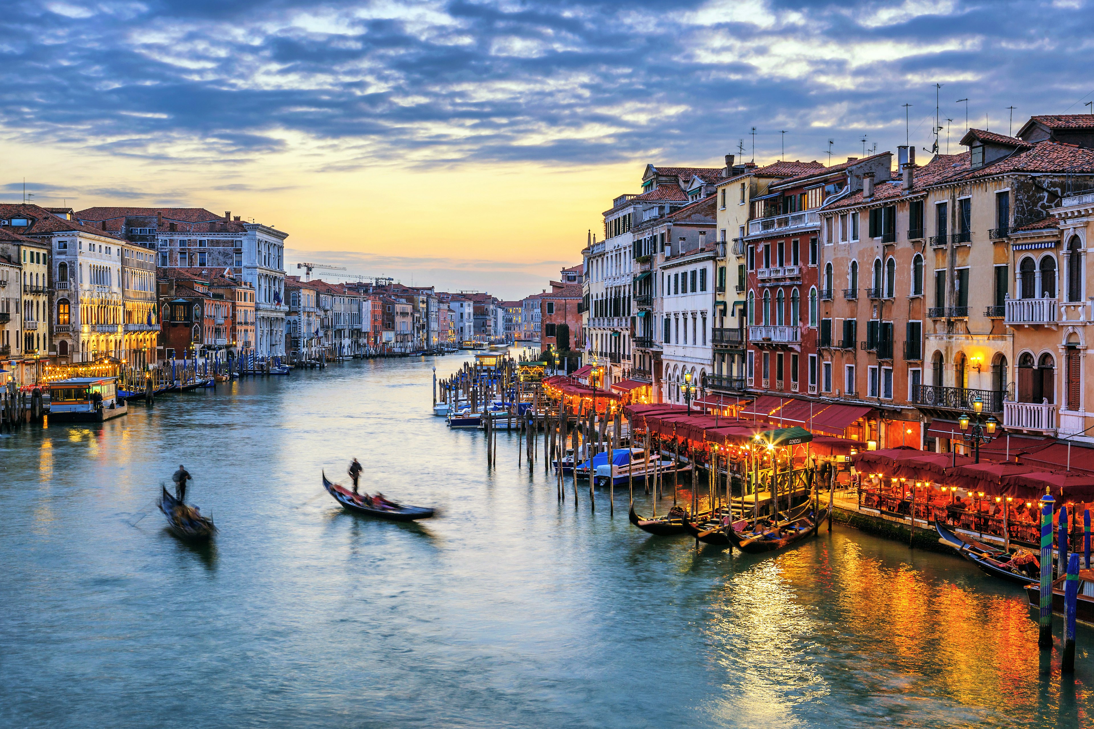 The Grand Canal in Venice at dusk, with restaurants lining the banks of the canal, and a gondolas silhouetted in the water