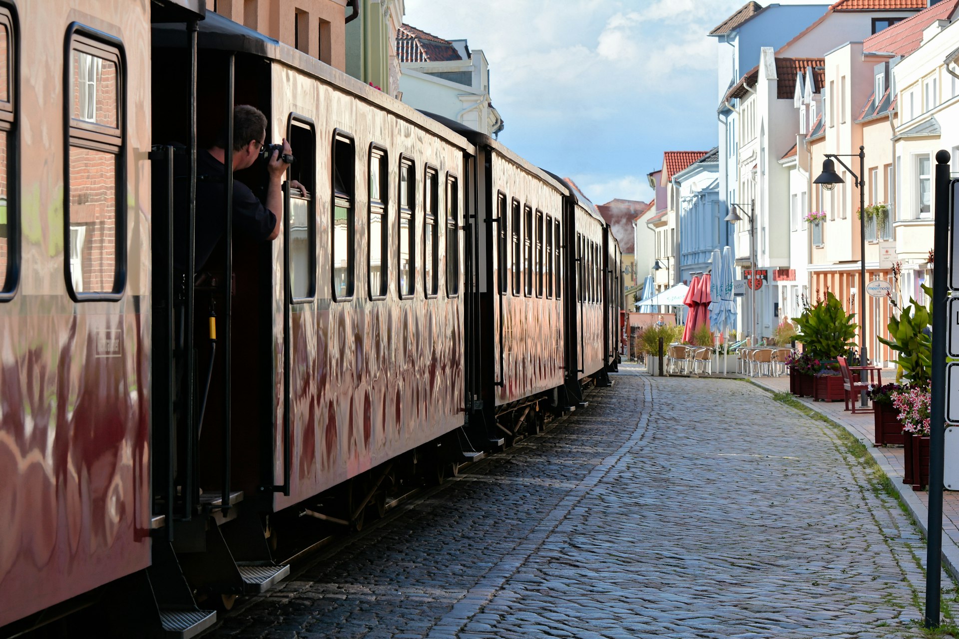 A man slightly leans out of a train to take a photo of an old cobbled stone town 