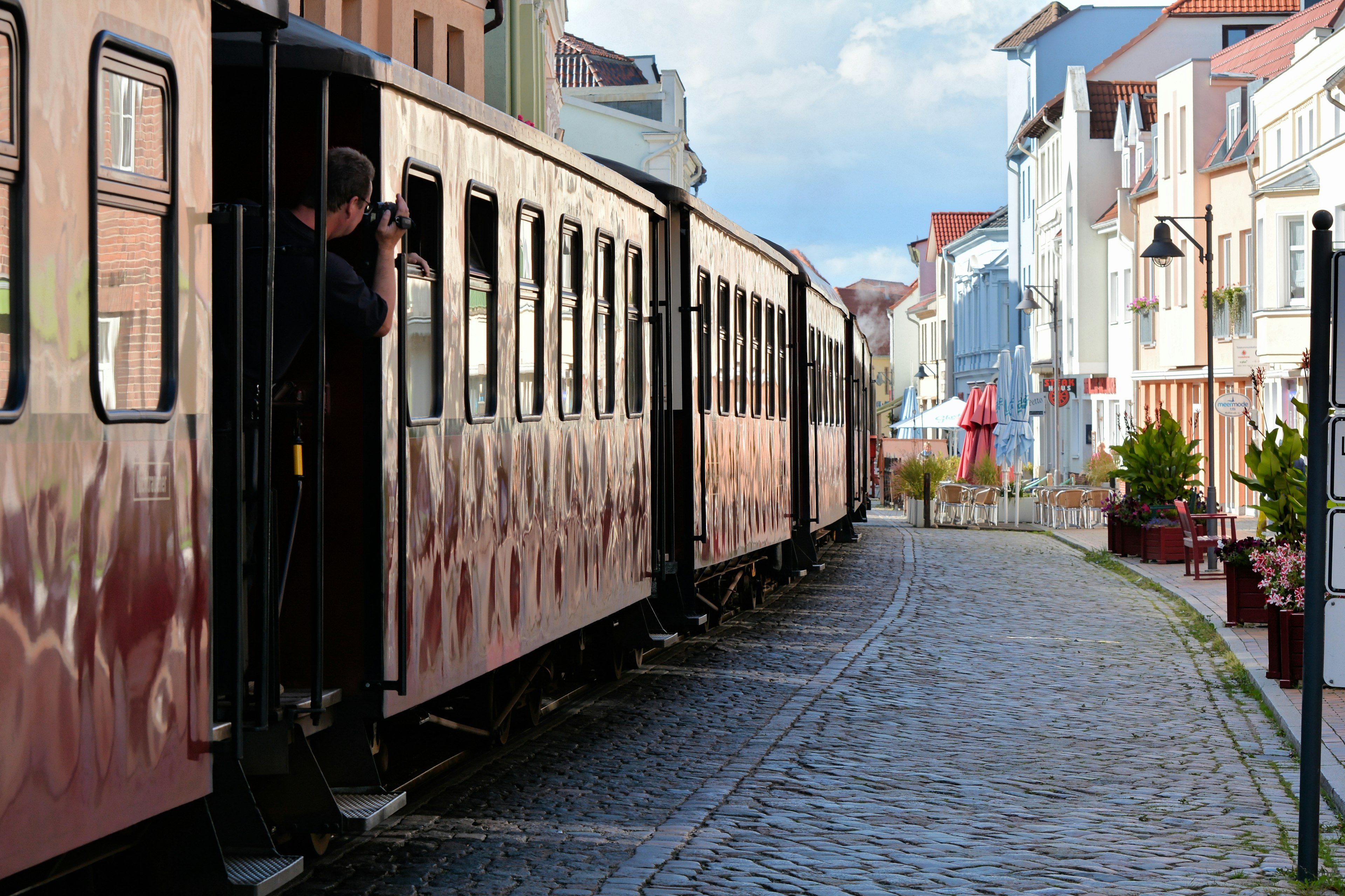 A man slightly leans out of a train to take a photo of an old cobbled stone town