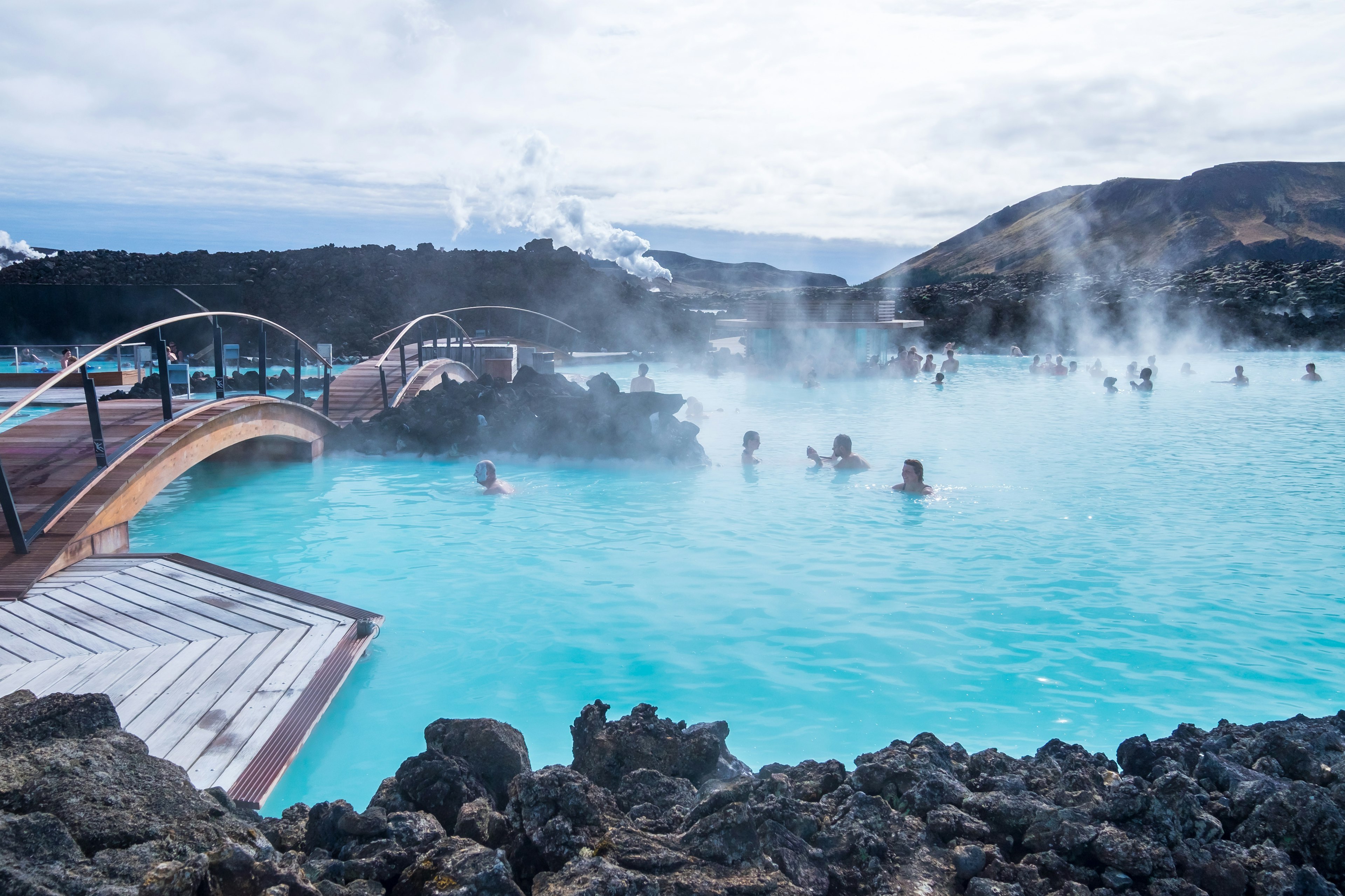 A wooden bridge crosses a lagoon filled with pale blue water. Steam is rising from the water and people are bathing in the water