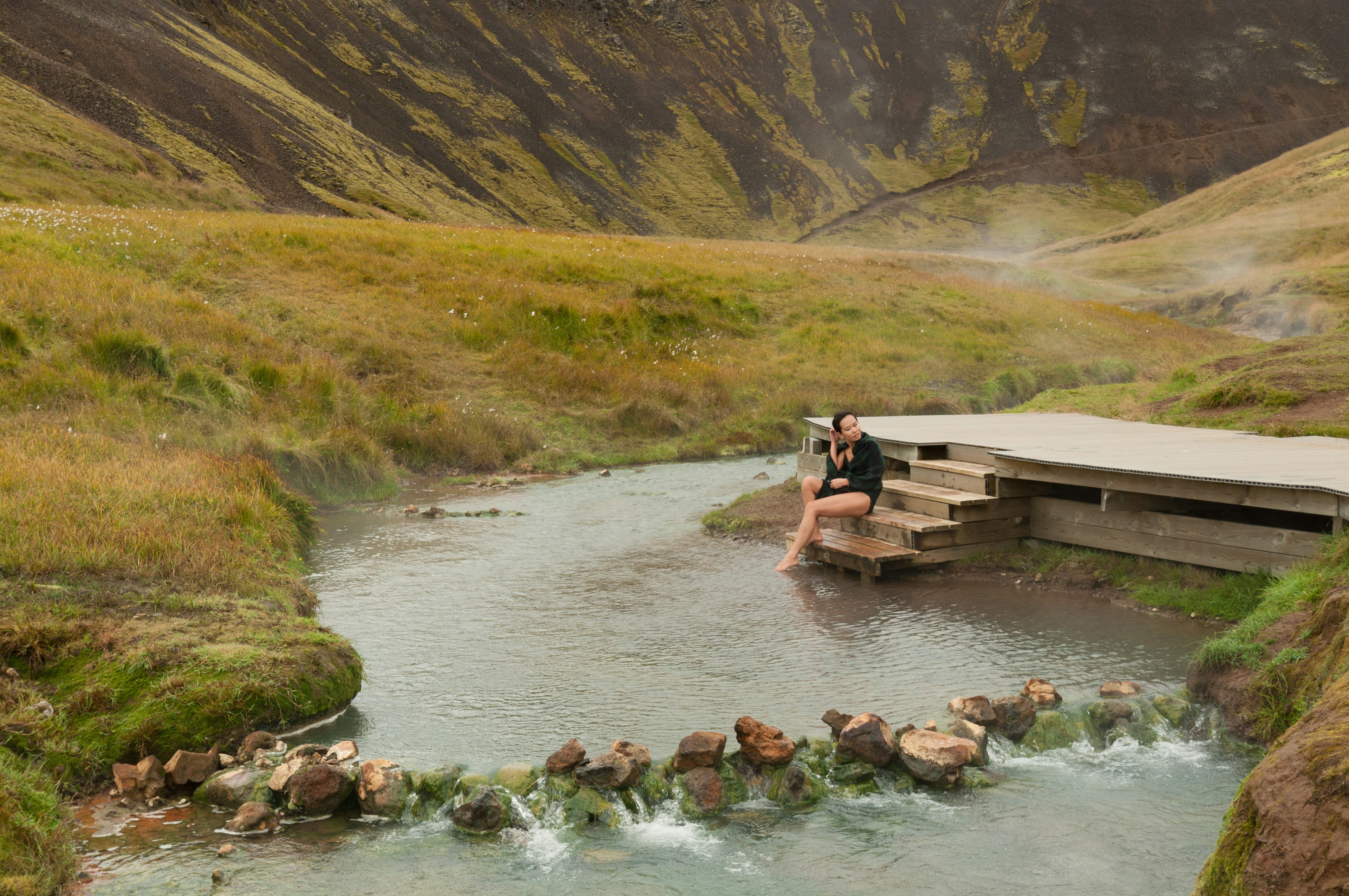 Woman sitting on wooden decking by a steaming hot-spring river cutting through the green landscape