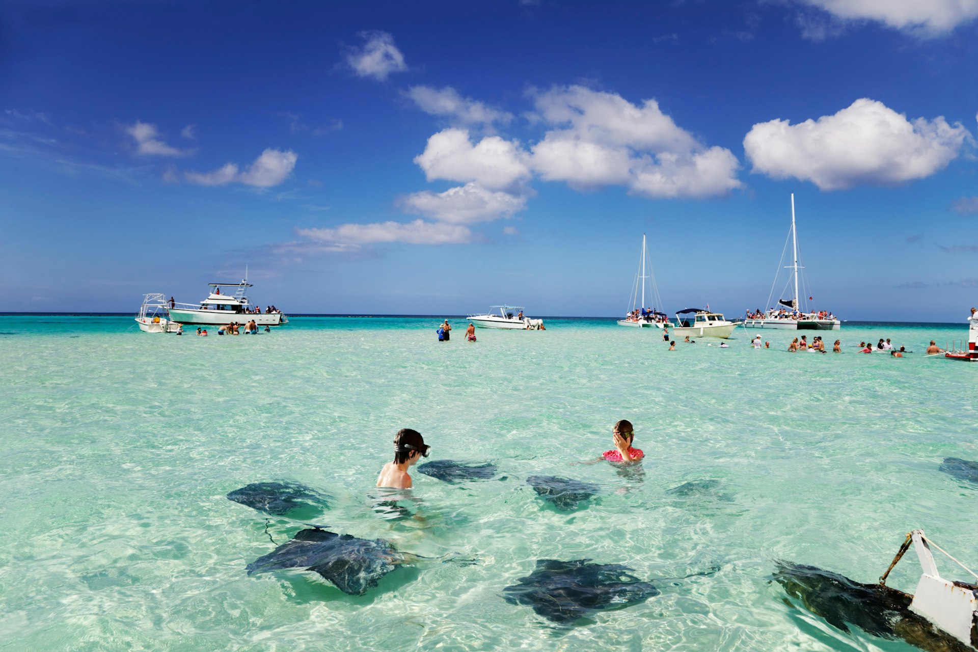 Two snorkelers pictured swimming with stingrays and sailboats in the distance in Stingray City, Grand Cayman