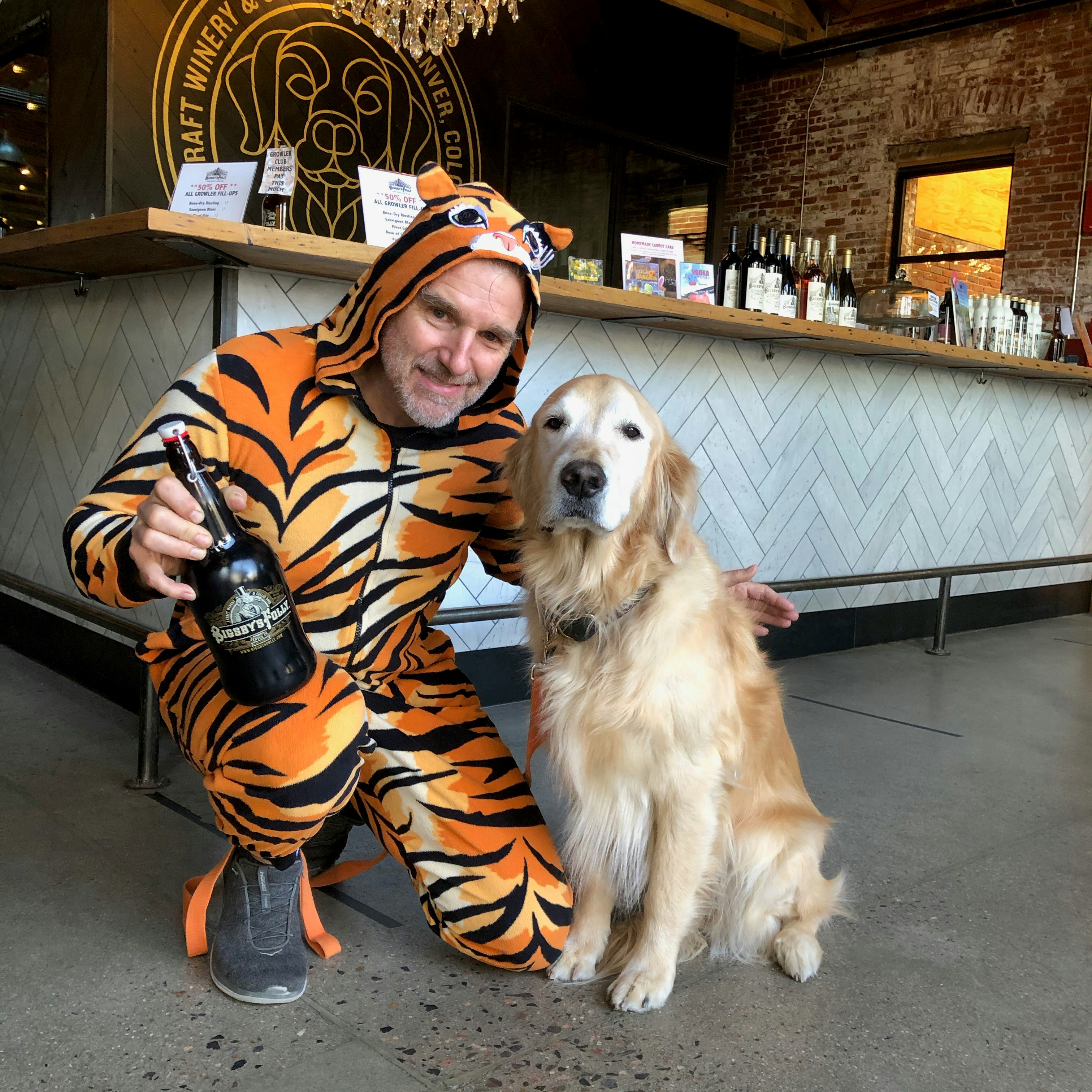 Man dressed in tiger costume crouches with his dog and a growler of wine