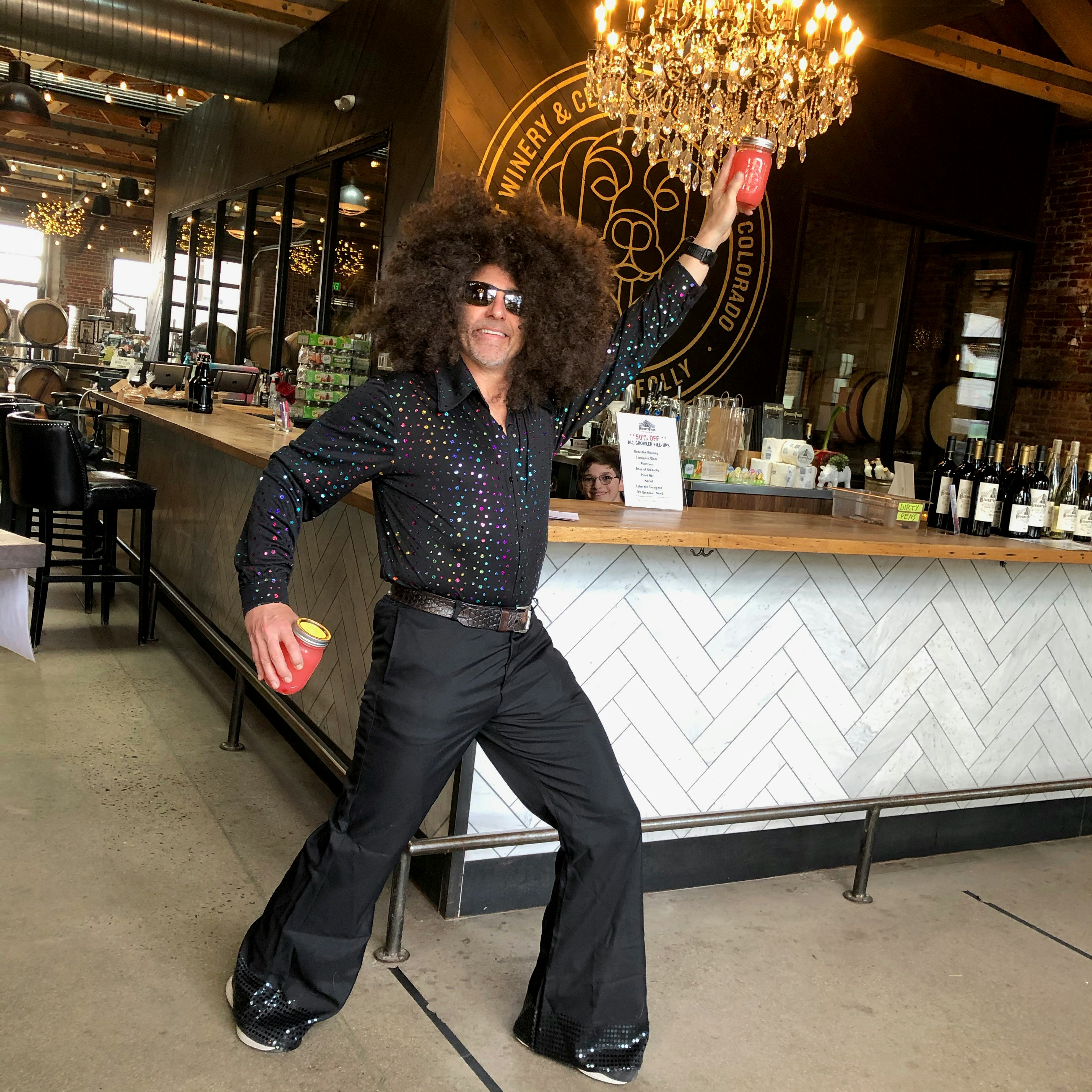 man dresses in black bellbottoms and sparkly top and dances in front of a bar