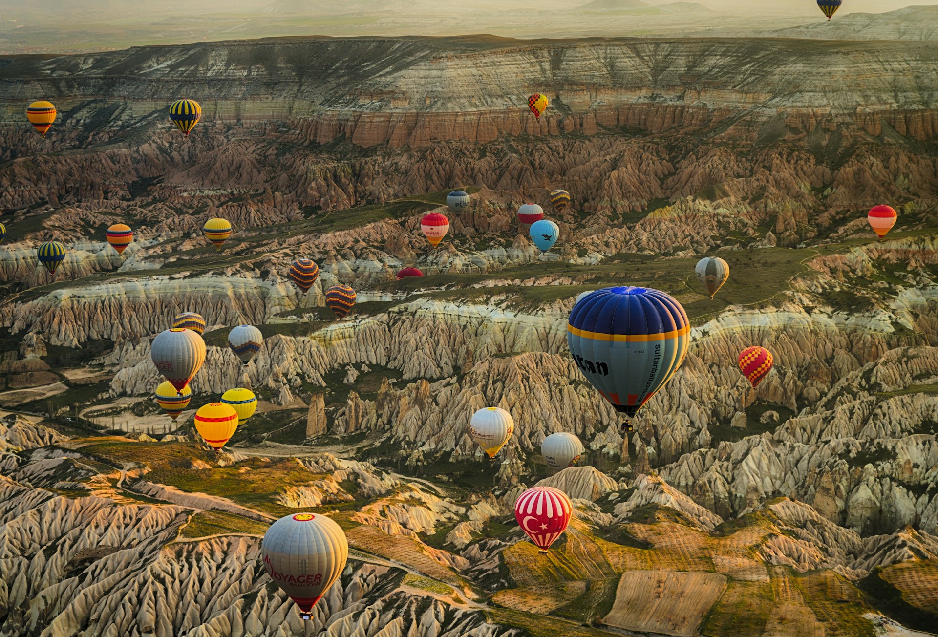 Aerial shot of brightly colored hot air balloons over a landscape with a lot of hills, exposed rock, and other geological features
