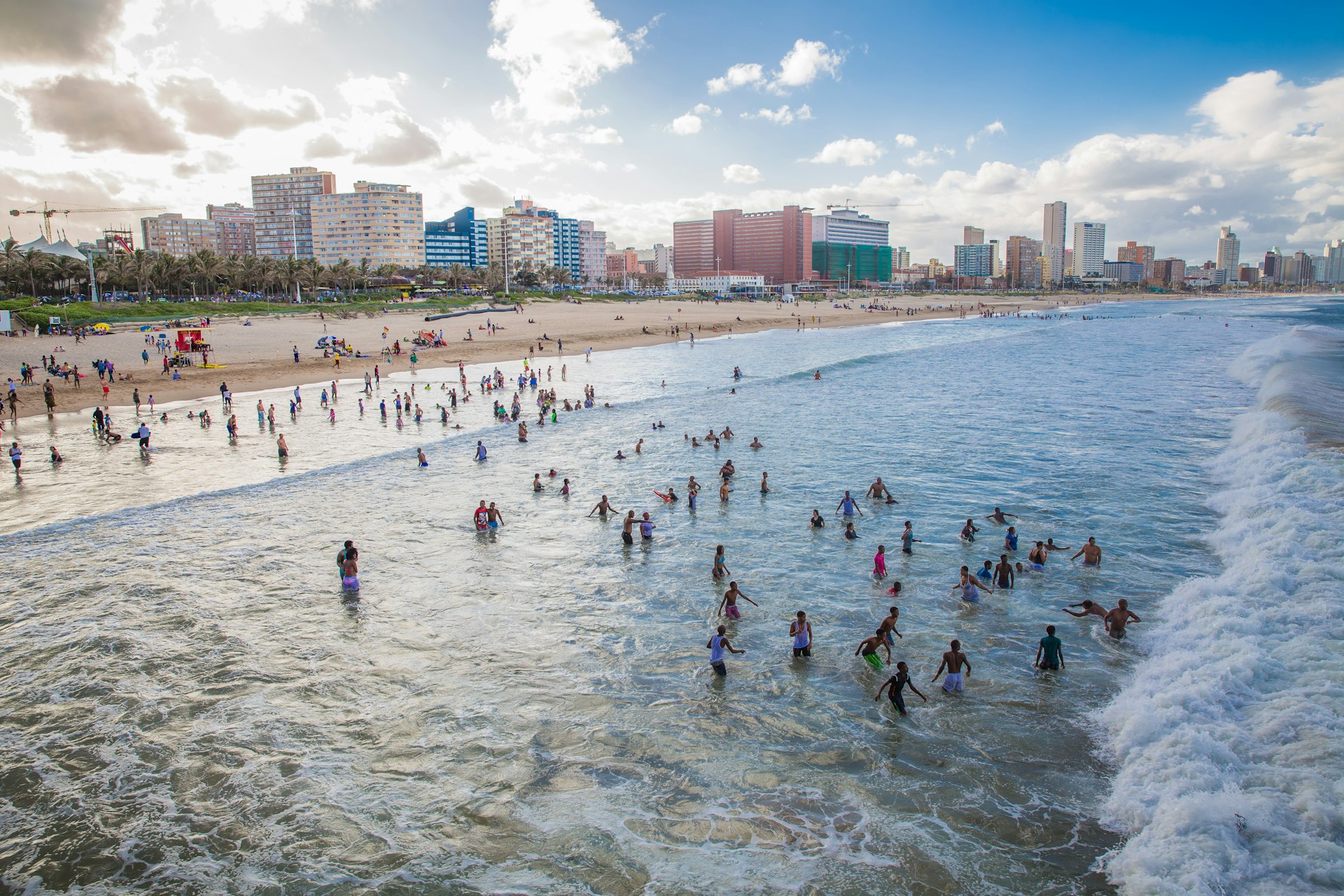 En flygfoto över stranden nära Durban, Sydafrika, där människor leker i vågorna.  I bakgrunden syns några av stadens skyskrapor.