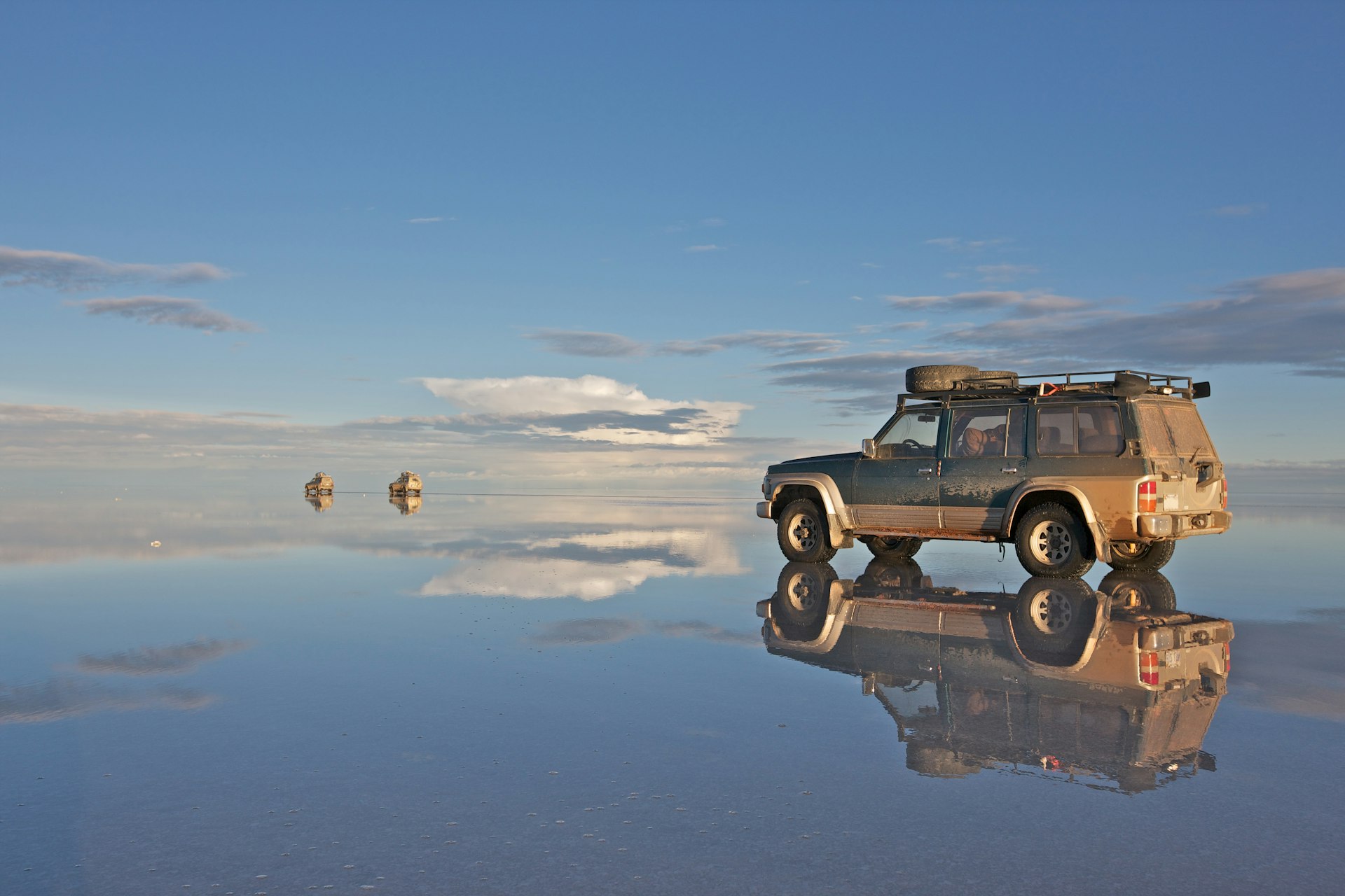 A 4WD stands in shallow water creating a near-perfect reflection. It is in an extensive flat area that seems to stretch endlessly