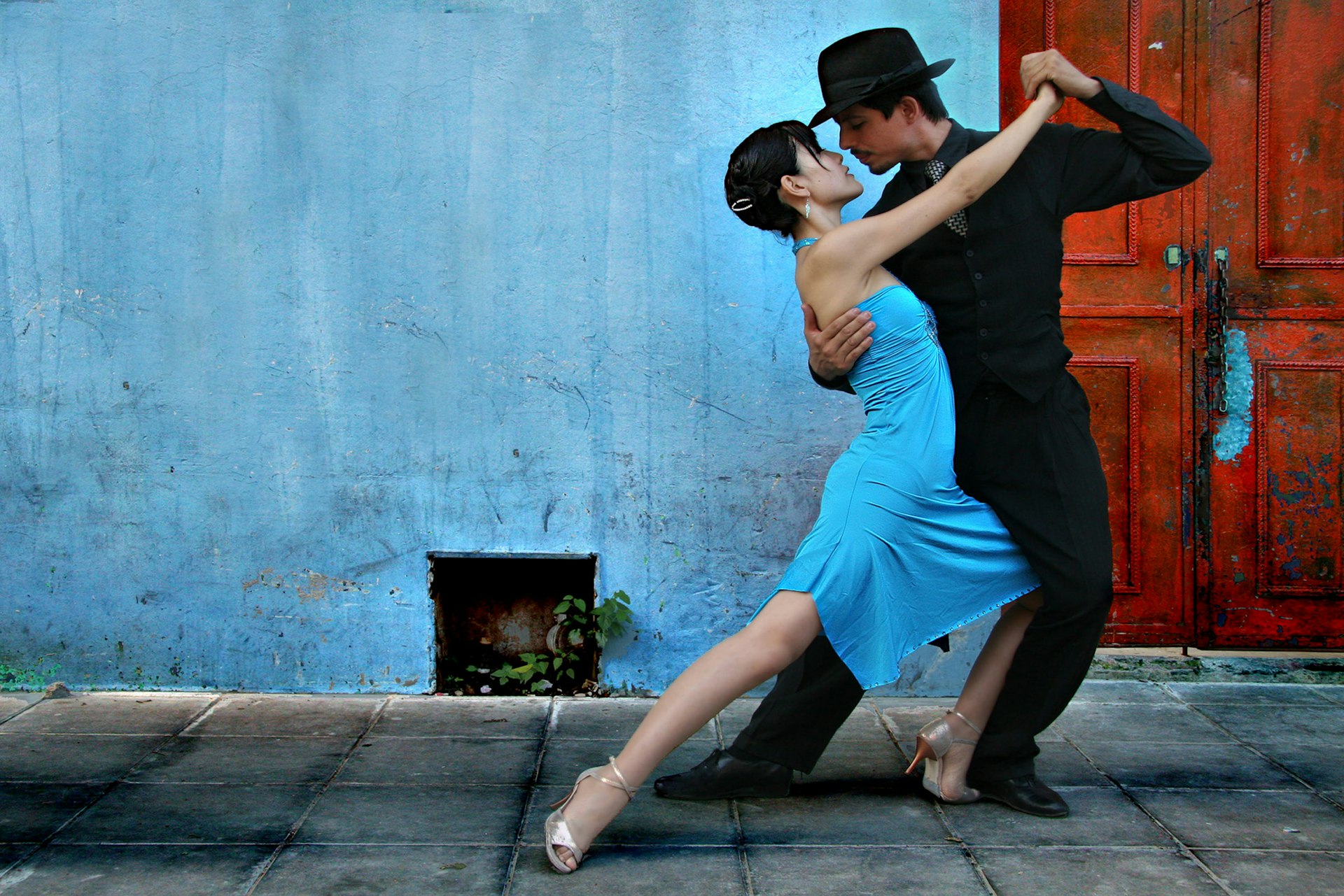 Two tango dancers pose together in front of a faded blue wall.