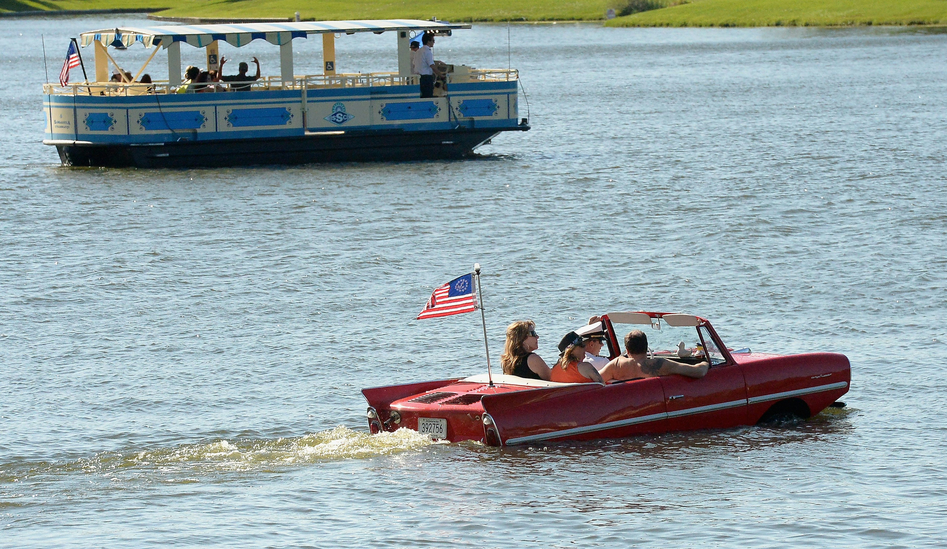 A group of people ride in a body of water in a red amphicar. There is a smaller boat in the the background.