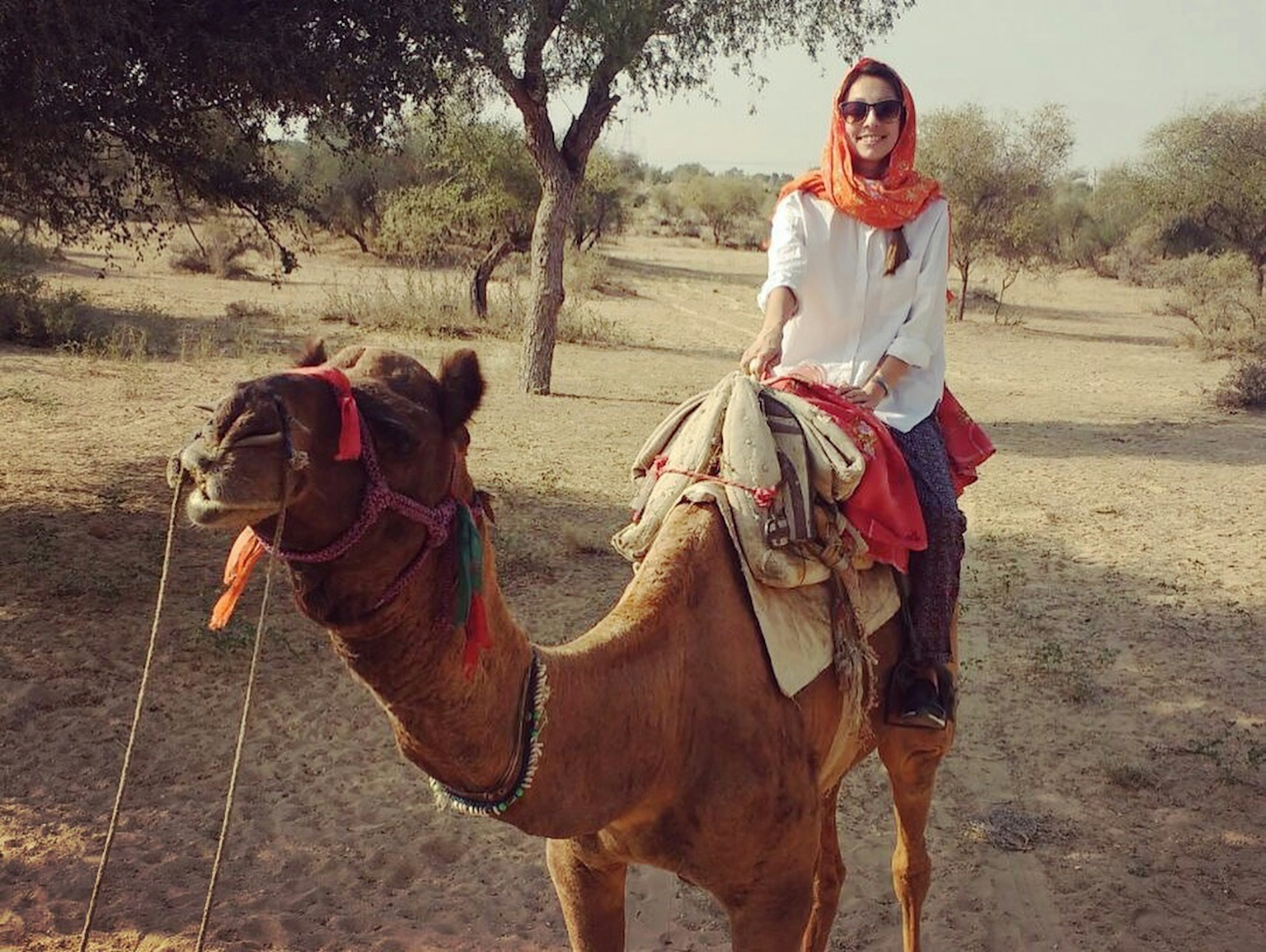 A woman smiles at the camera from the back of a camel in a dusty landscape