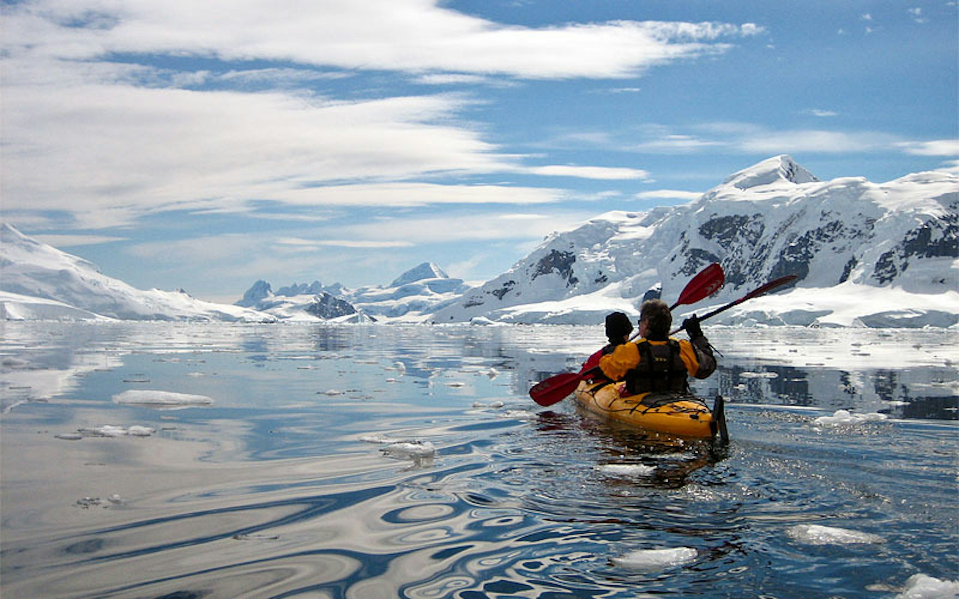Two people in a kayak move through icy water, surrounded by snowy hills and ridges