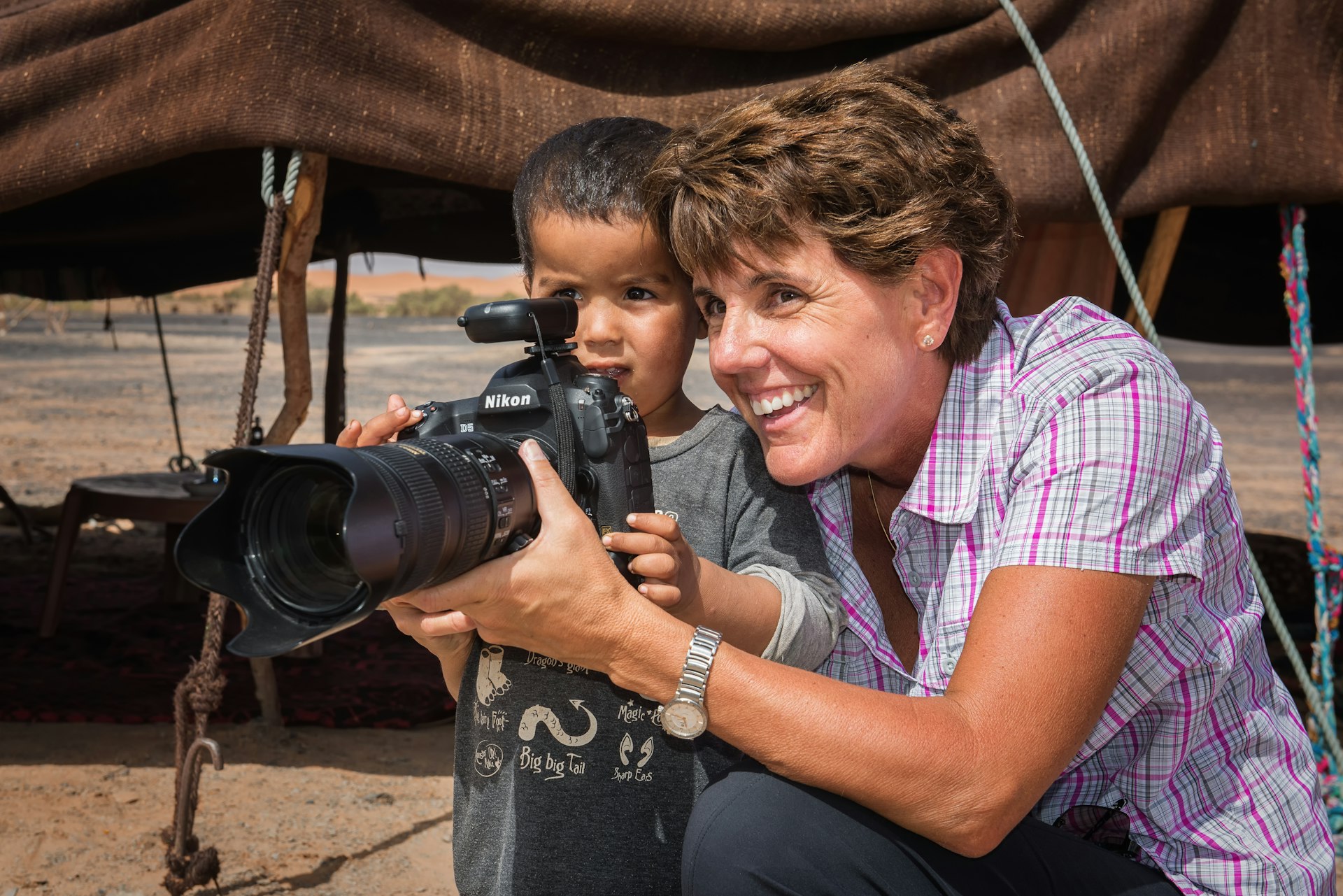 A woman shows a young child how to operate a camera. They're in a desert area with a canopy behind them propped up on sticks