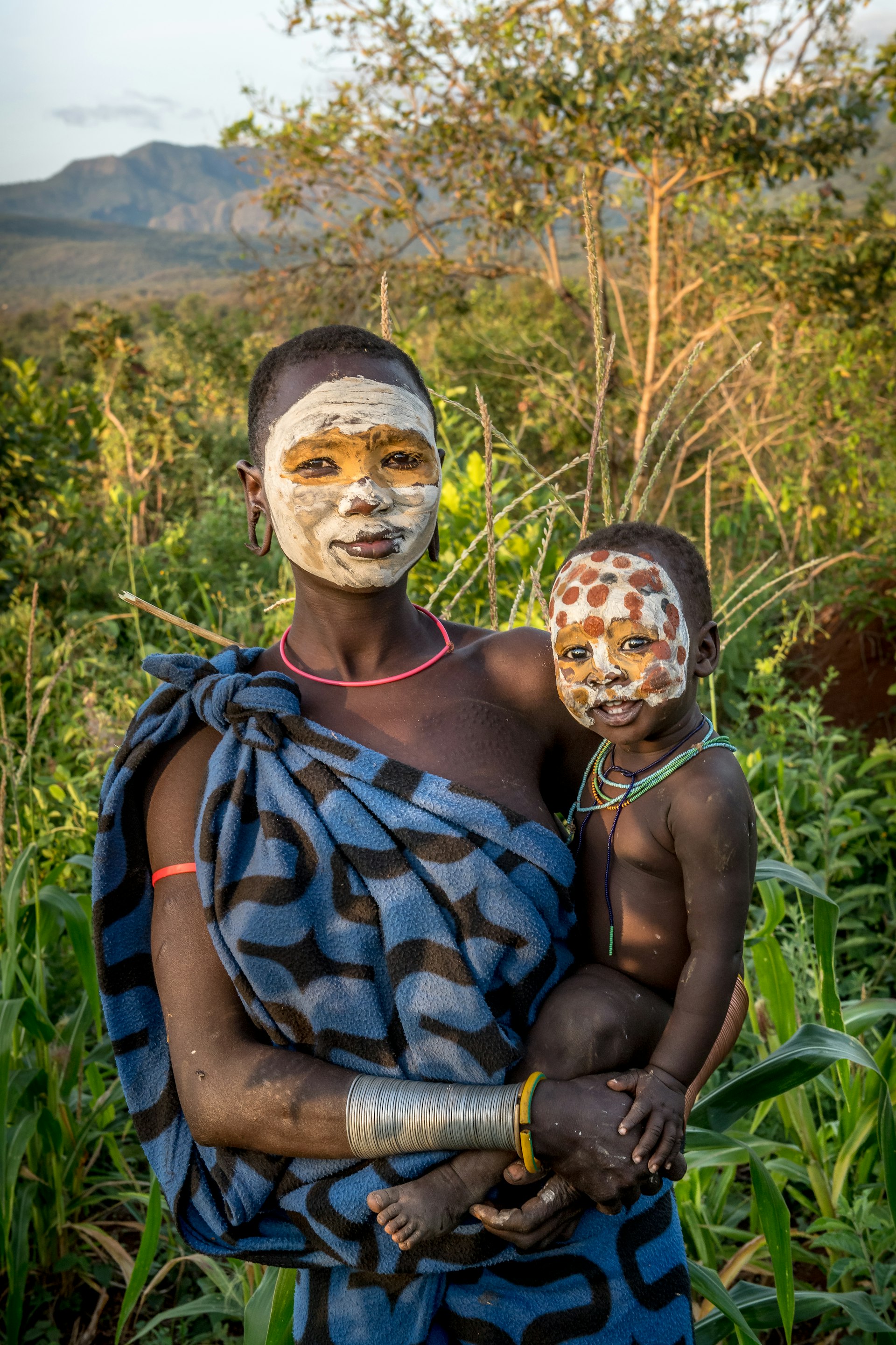 A woman holds a child. They both have their faces decorated with white and gold paint