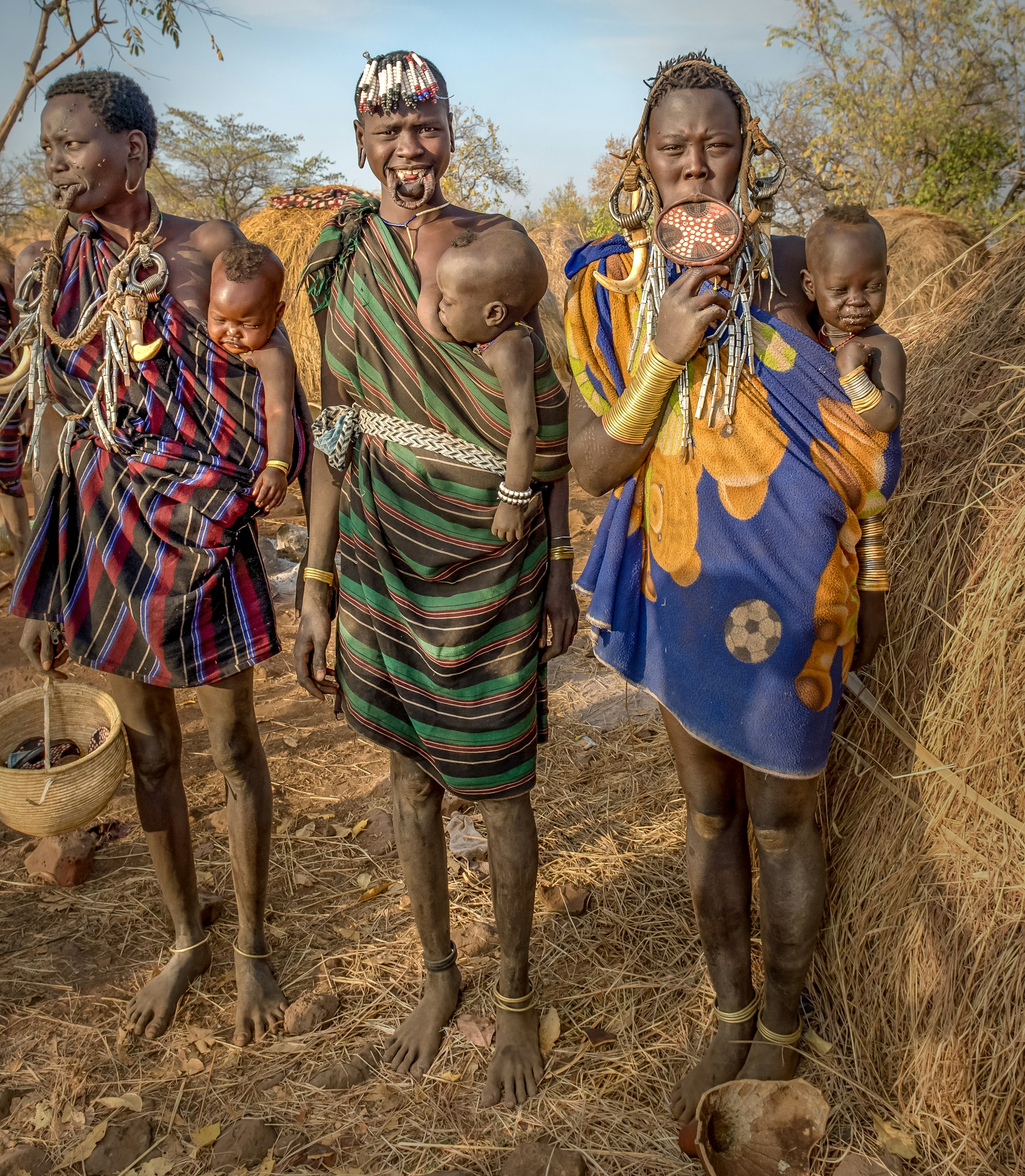 Three tribeswomen, each with a lip plate in their bottom lips, stand together with three infants held in slings across their hips