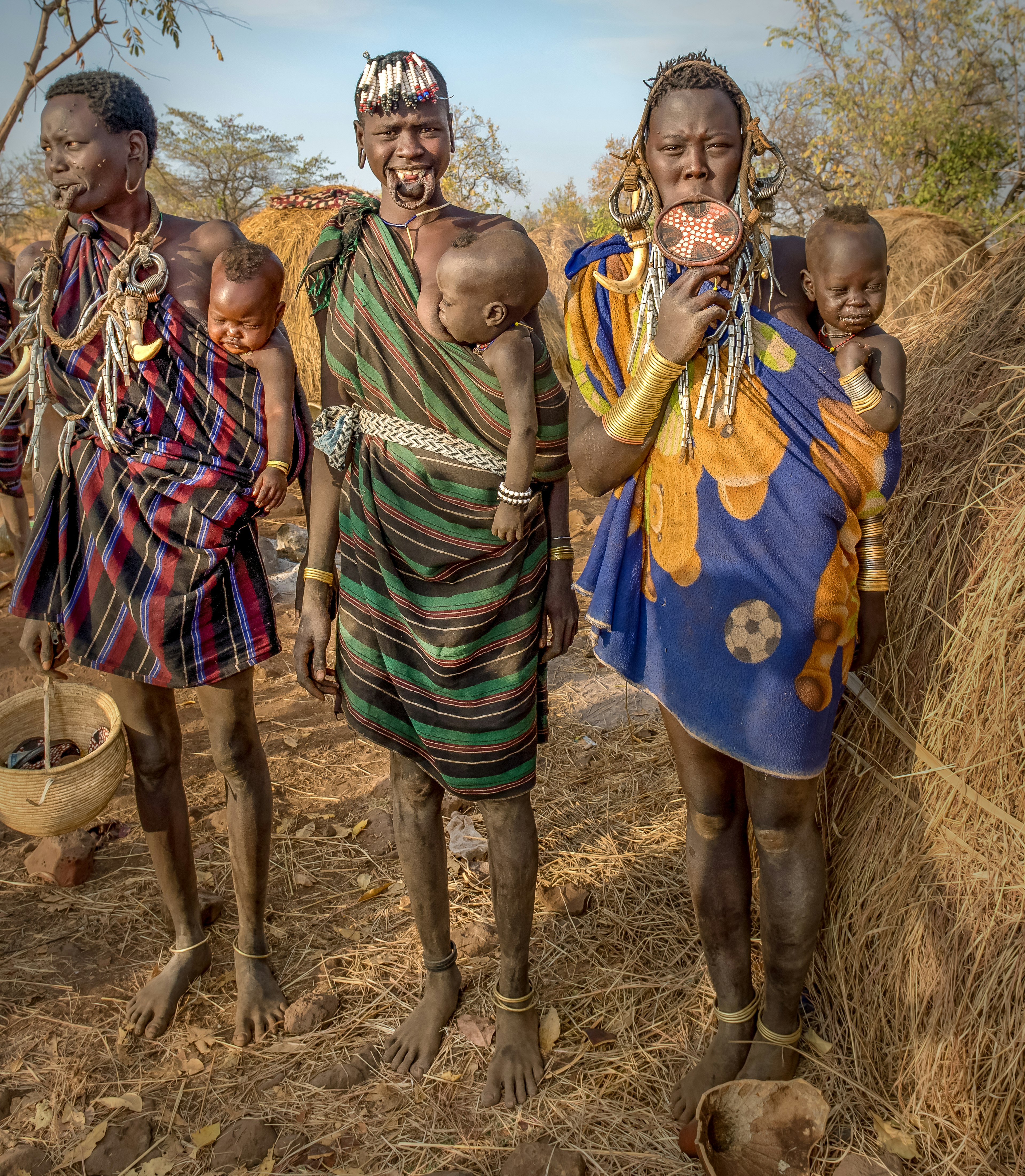 Three tribeswomen, each with a lip plate in their bottom lips, stand together with three infants held in slings across their hips