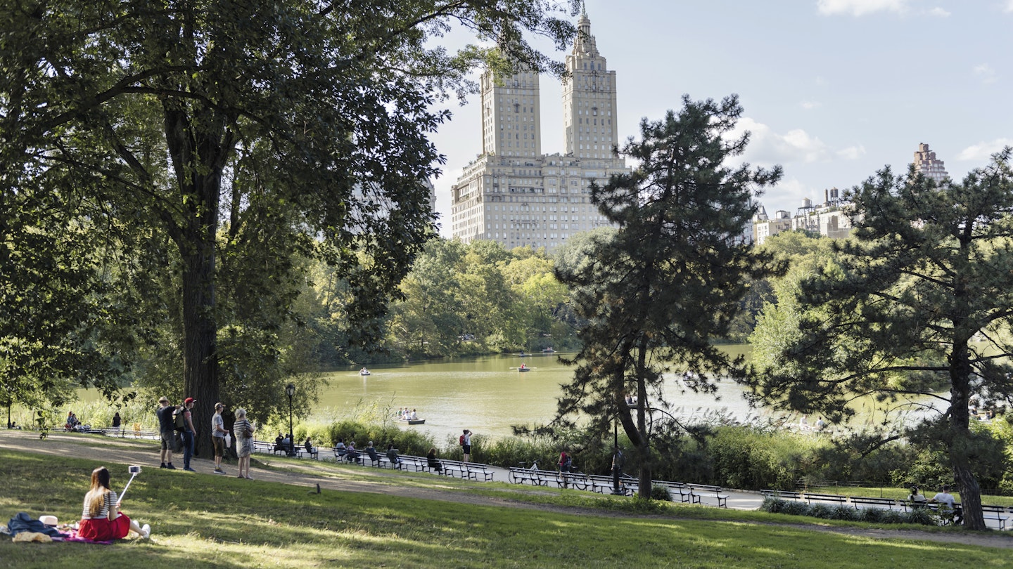 People sit around The Lake in Central Park on a sunny afternoon. The San Remo apartment building can be seen in the distance.