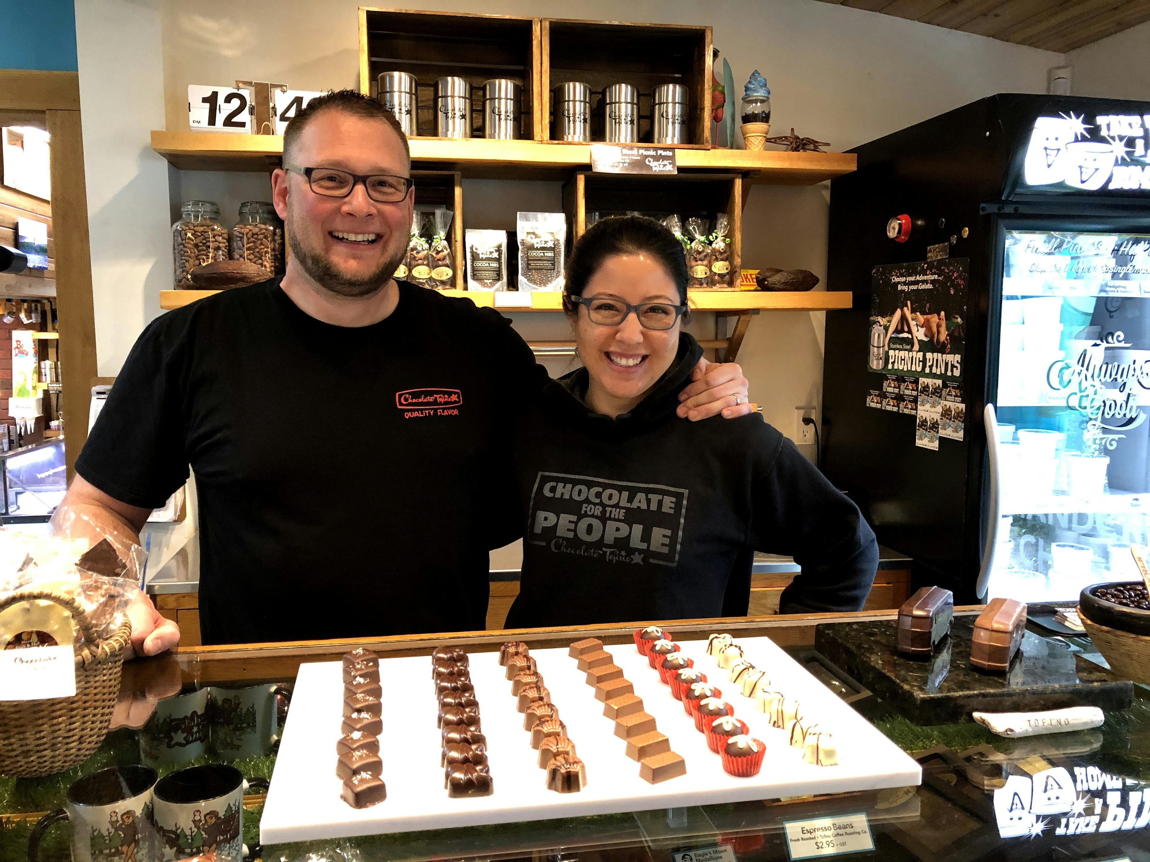 A man and a woman stand together smiling at the camera. In front of them is a tray of artisanal chocolates