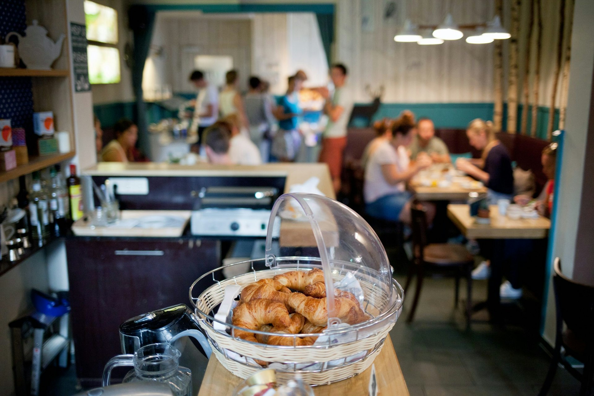 A close-up of some croissants on a bar in a half-domed glass bowl; the blurred background shows people sitting in a communal area of a hostel