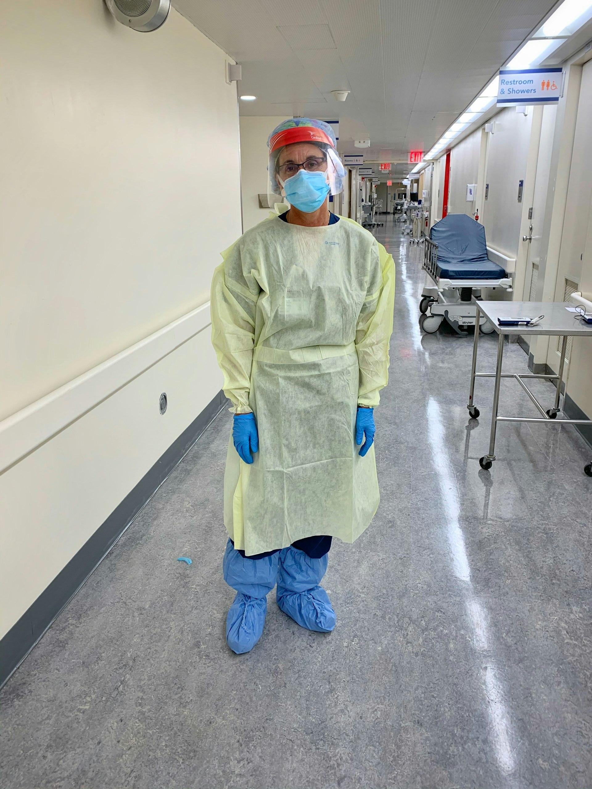 A person in full protective equipment, including a full-body gown, plastic boots over shoes, gloves, face mask and plastic visor, stands in a hospital corridor
