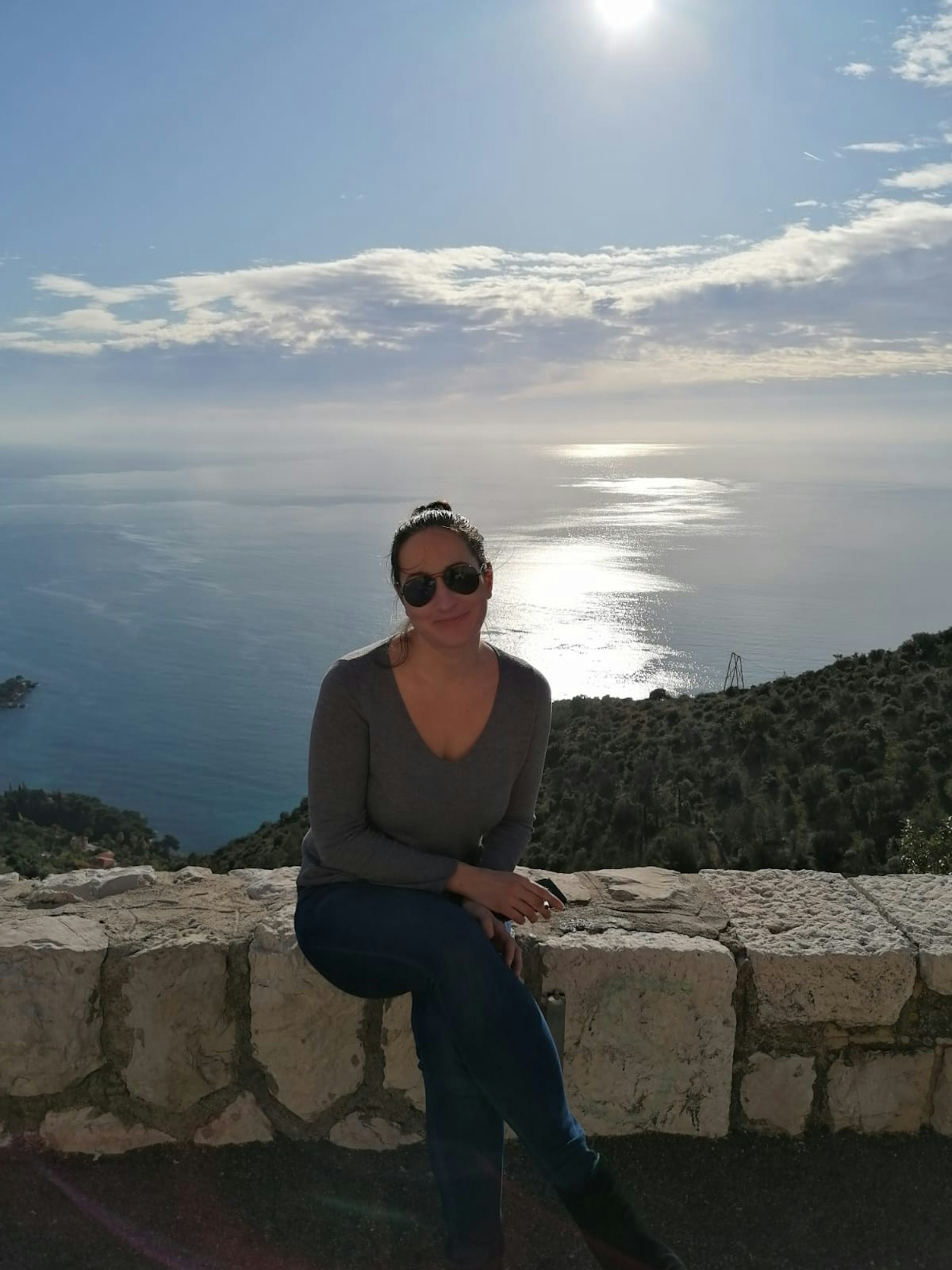A woman sitting on a wall wearing sunglasses. The sea is sparkling in the distance