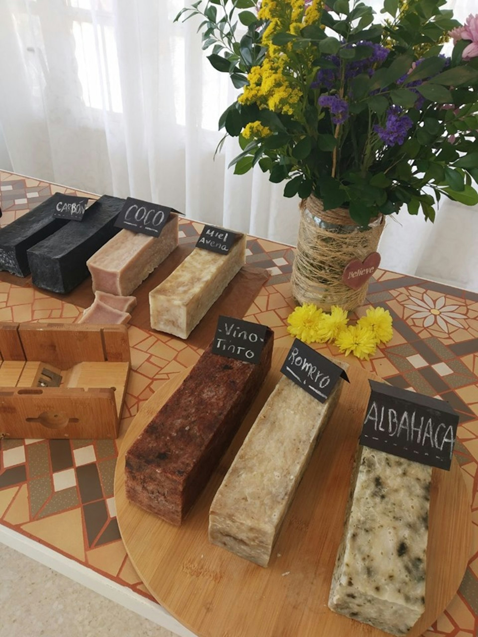 Large rectangular blocks of soap on a table, each with a label. A large vase of flowers stands behind