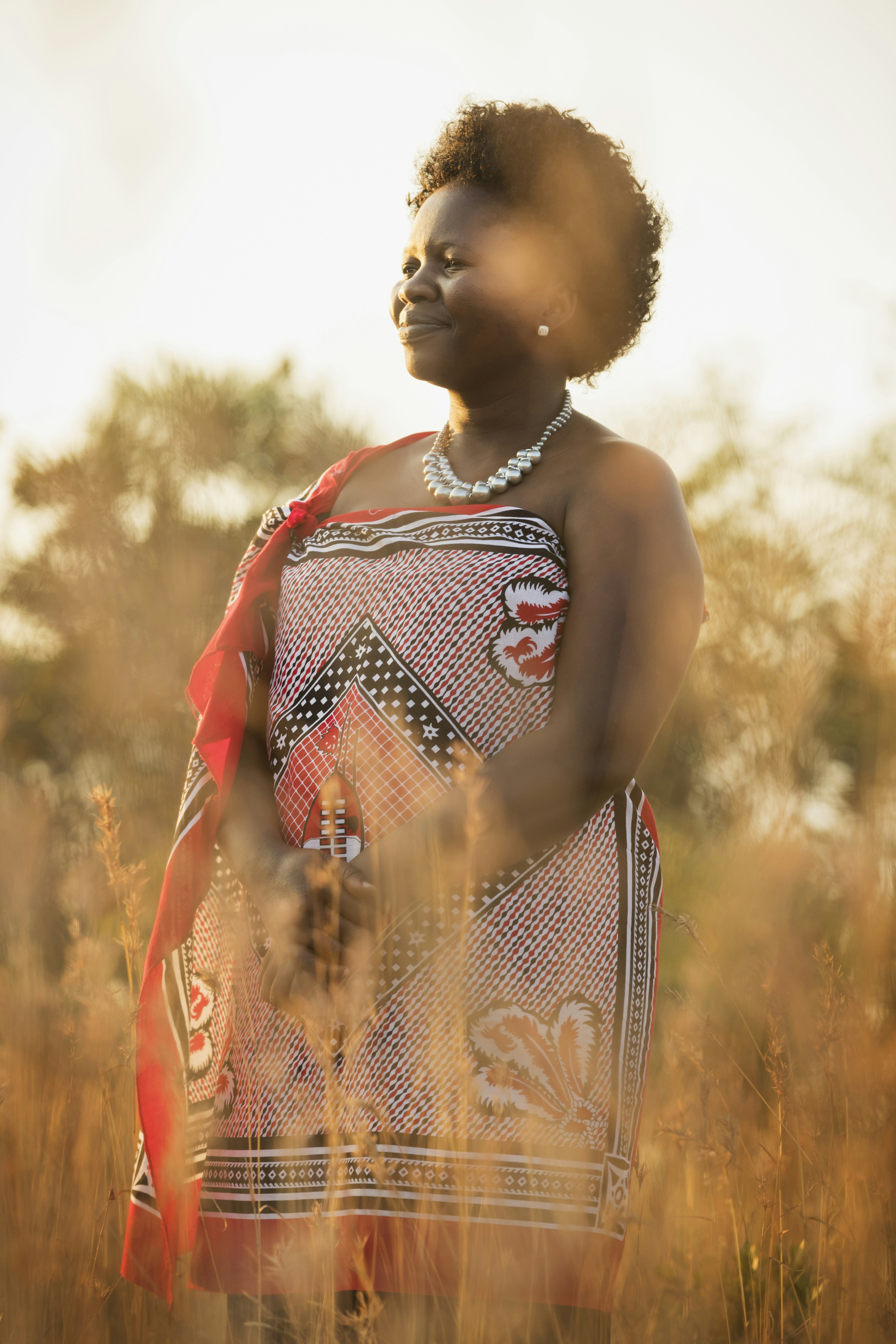 A woman, dressed in a traditional African patterned cloth of black and red, stands in long grass smiling