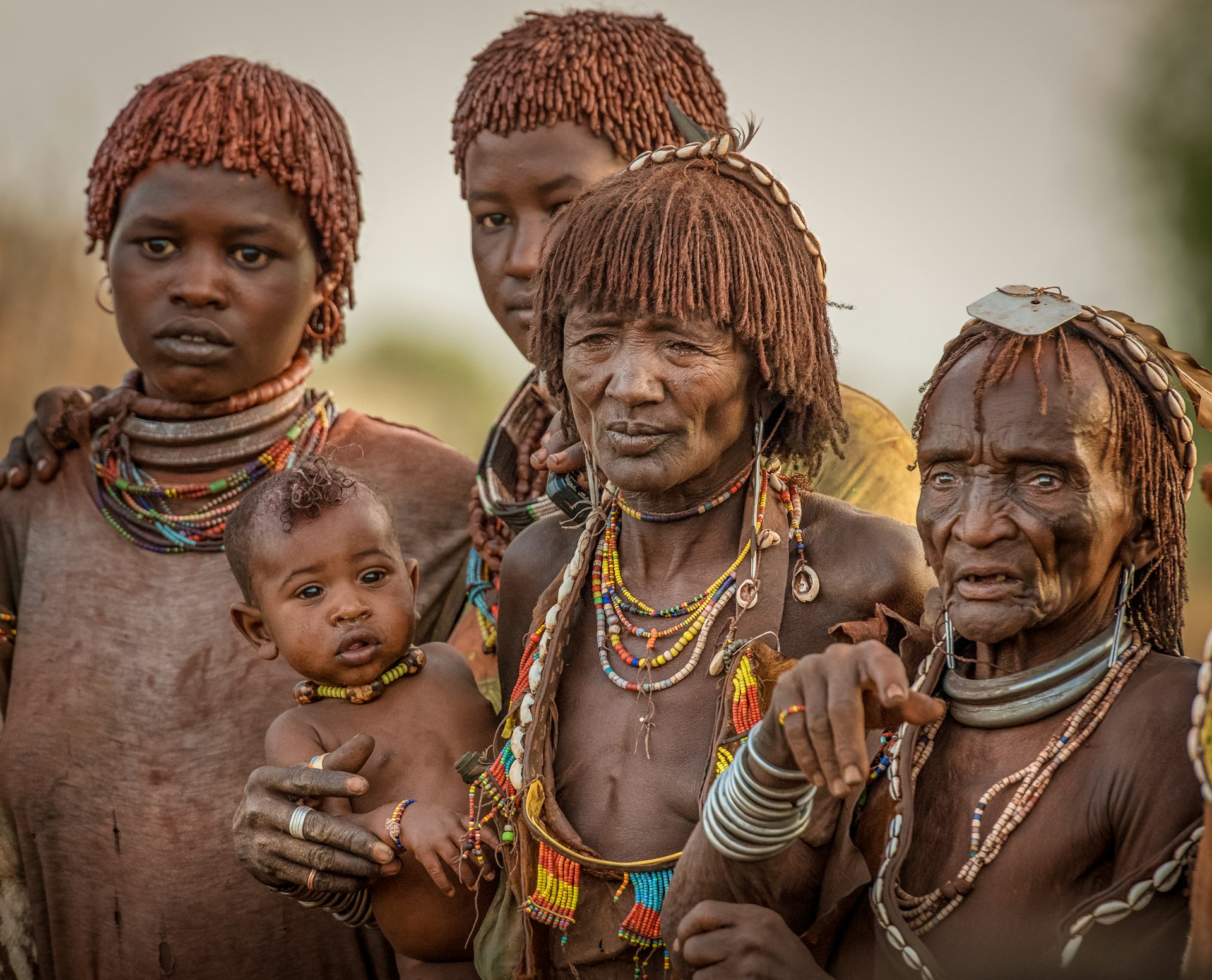 A group of tribes people of all ages stand together. The woman in the center is holding a young baby
