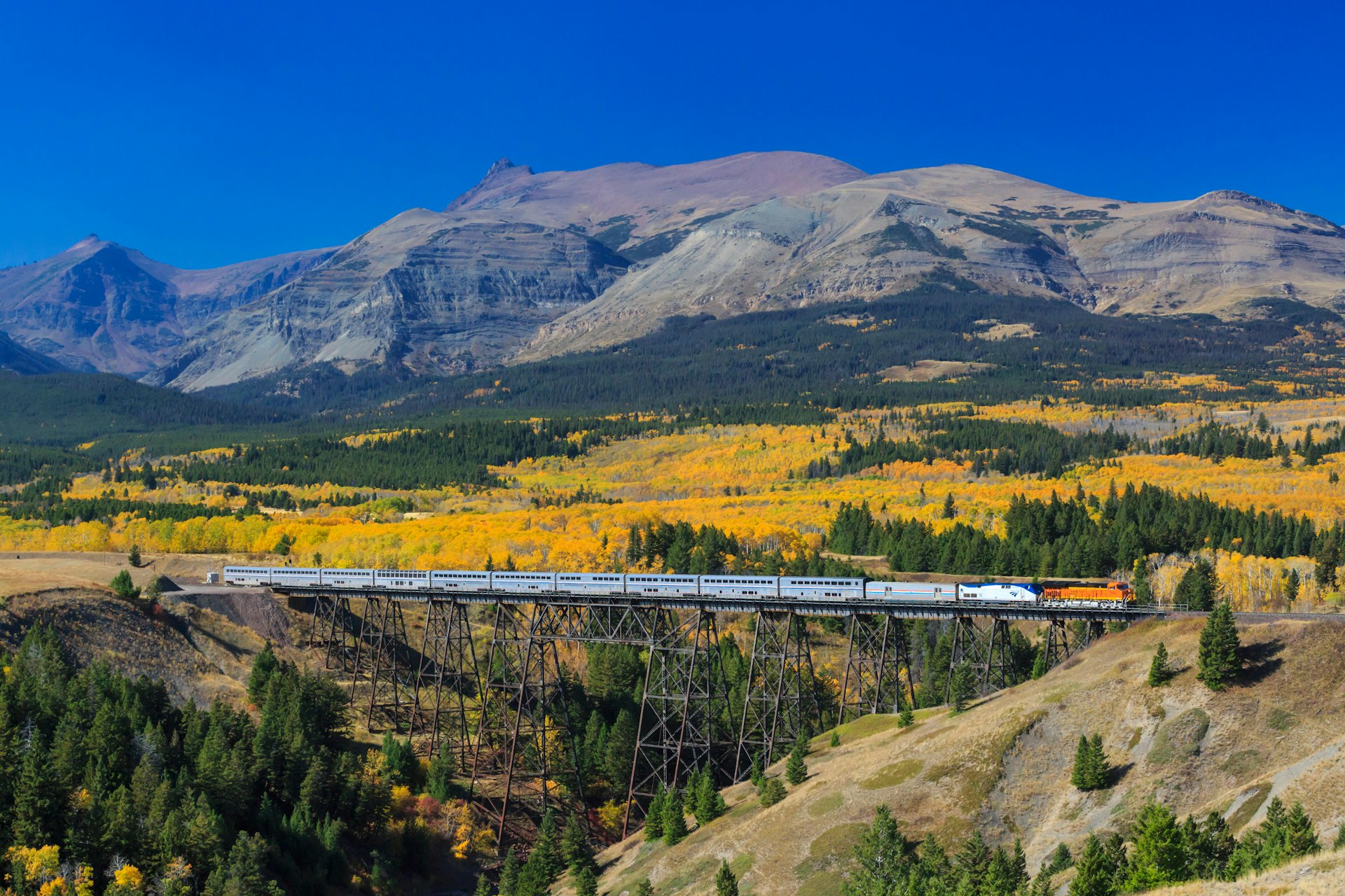 passenger train crossing over trestle in autumn below peaks of glacier national park near east glacier park, montana. Image shot 10/2015. Exact date unknown.