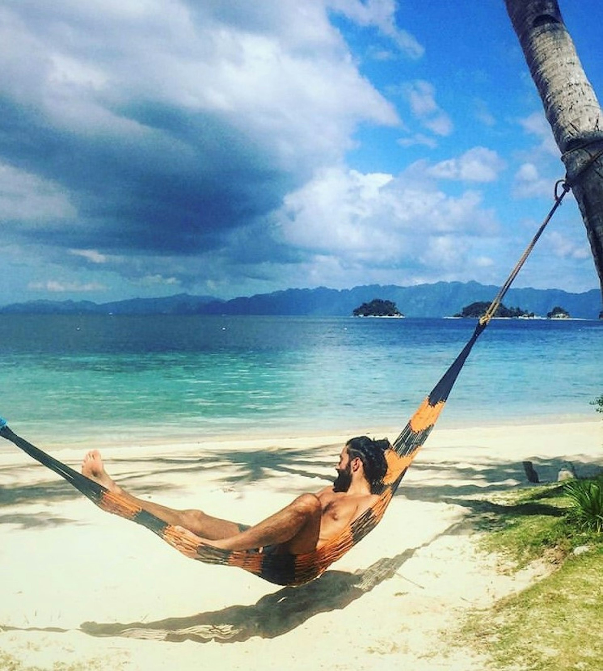 A man lies in a hammock slung between palm trees. He is looking out at the clear, calm, turquoise sea