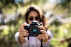 A brunette woman wearing sunglasses holds a DSLR camera in front of her.