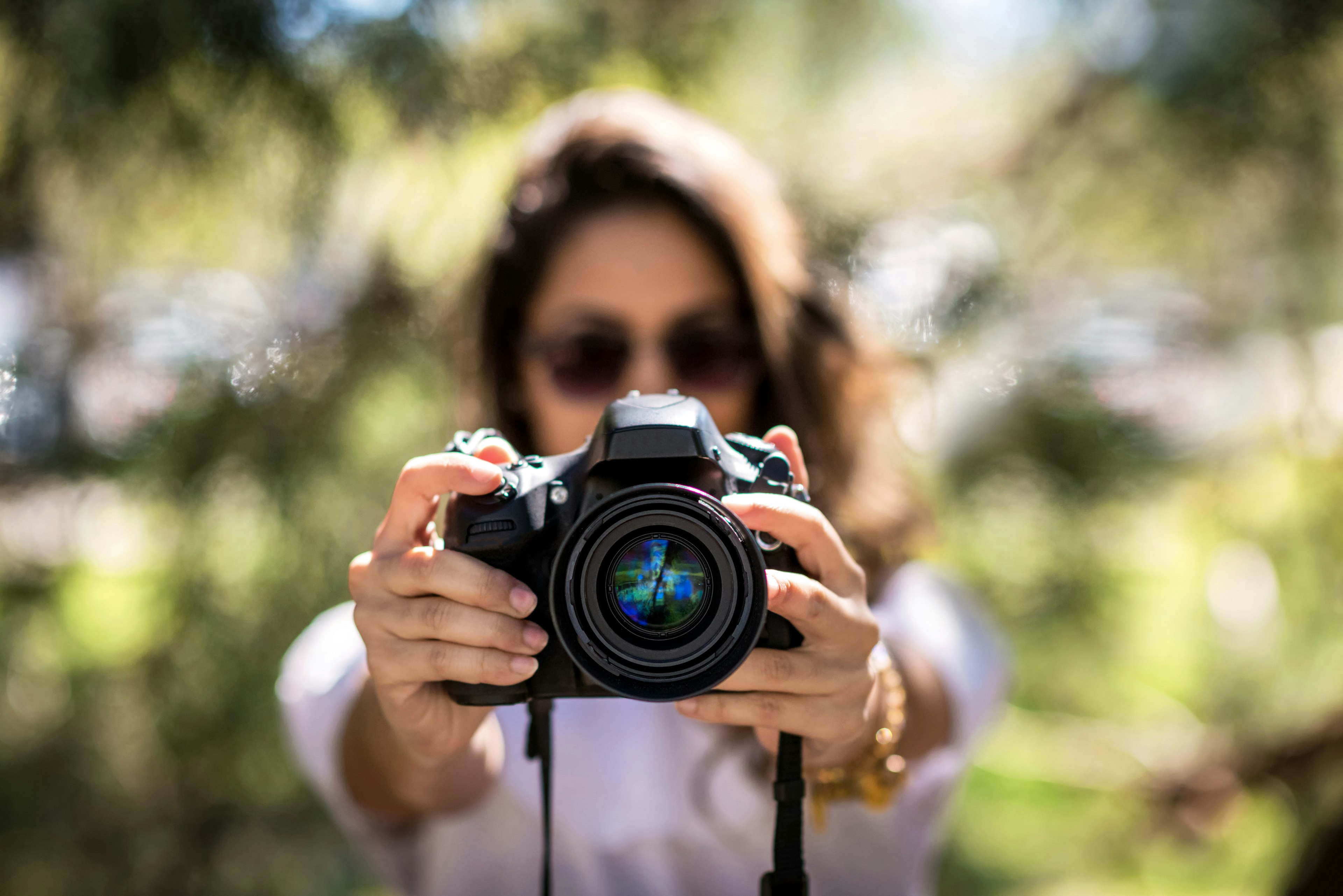 A brunette woman wearing sunglasses holds a DSLR camera in front of her.