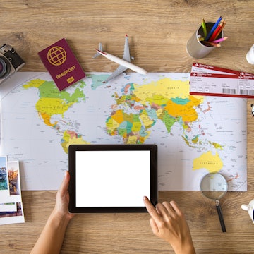 Hand holding a blank white screen smart phone, travel stuff on desktop.