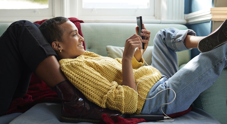 Smiling woman text messaging on smart phone while leaning on friend's leg in living room