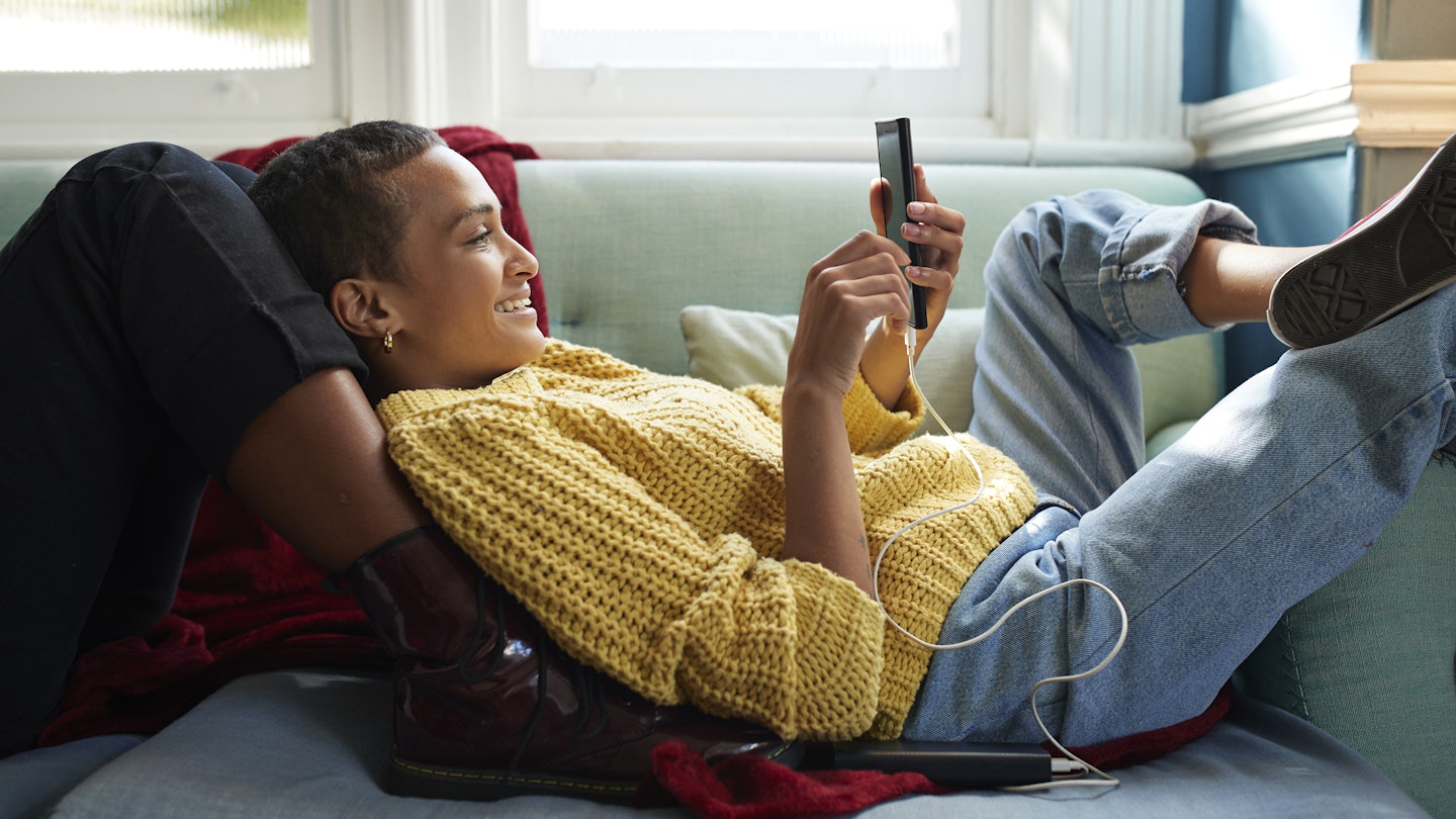 Smiling woman text messaging on smart phone while leaning on friend's leg in living room