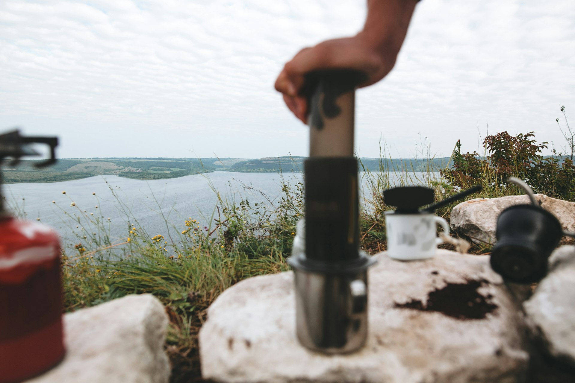 Focus on grass on cliff and blurred image of traveler pressing aeropress on metal mug on cliff at lake, brewing alternative coffee at camping. Making hot drink at picnic outdoors