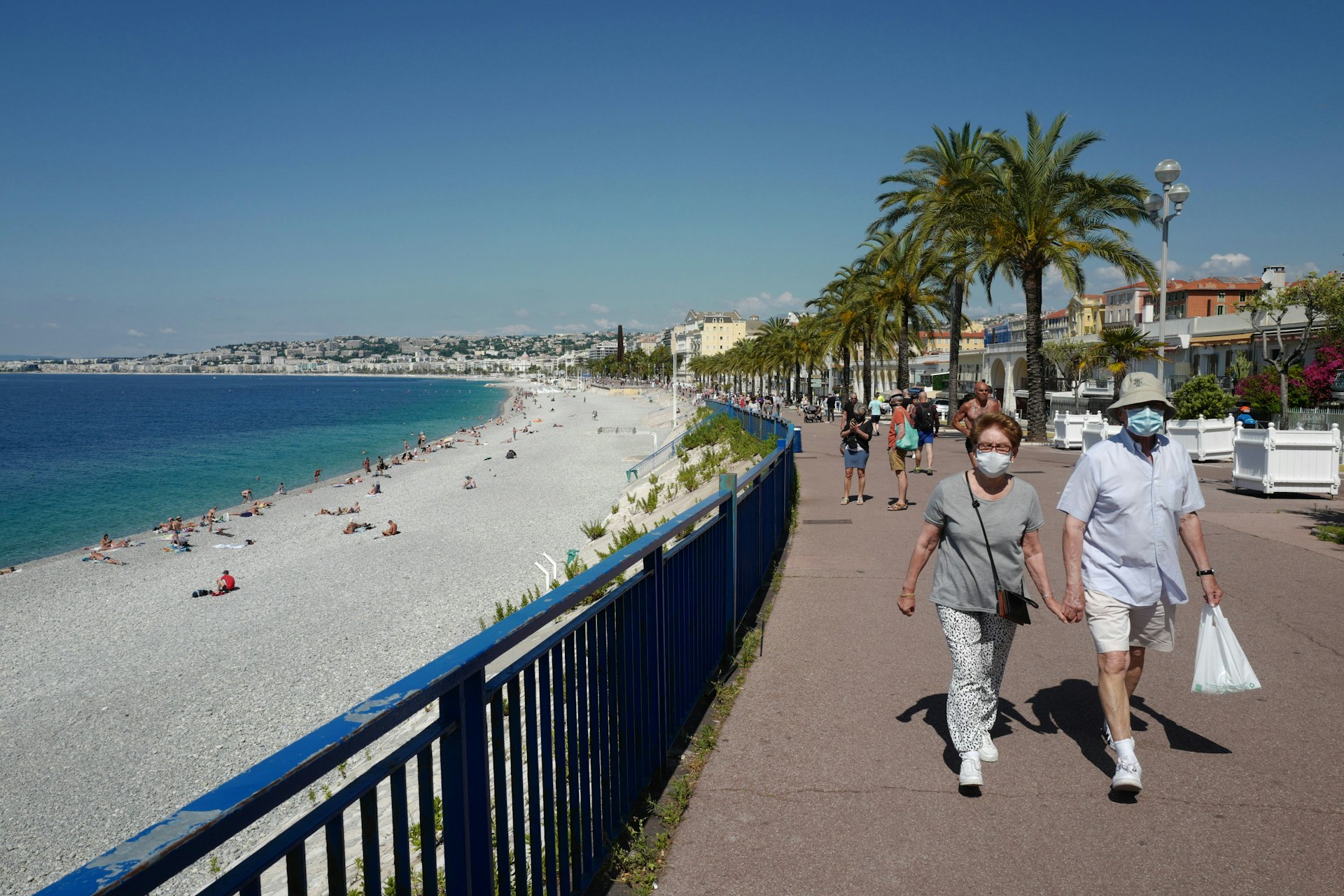 A couple wearing face masks stroll along a wide sidewalk beside the beach in Nice, France
