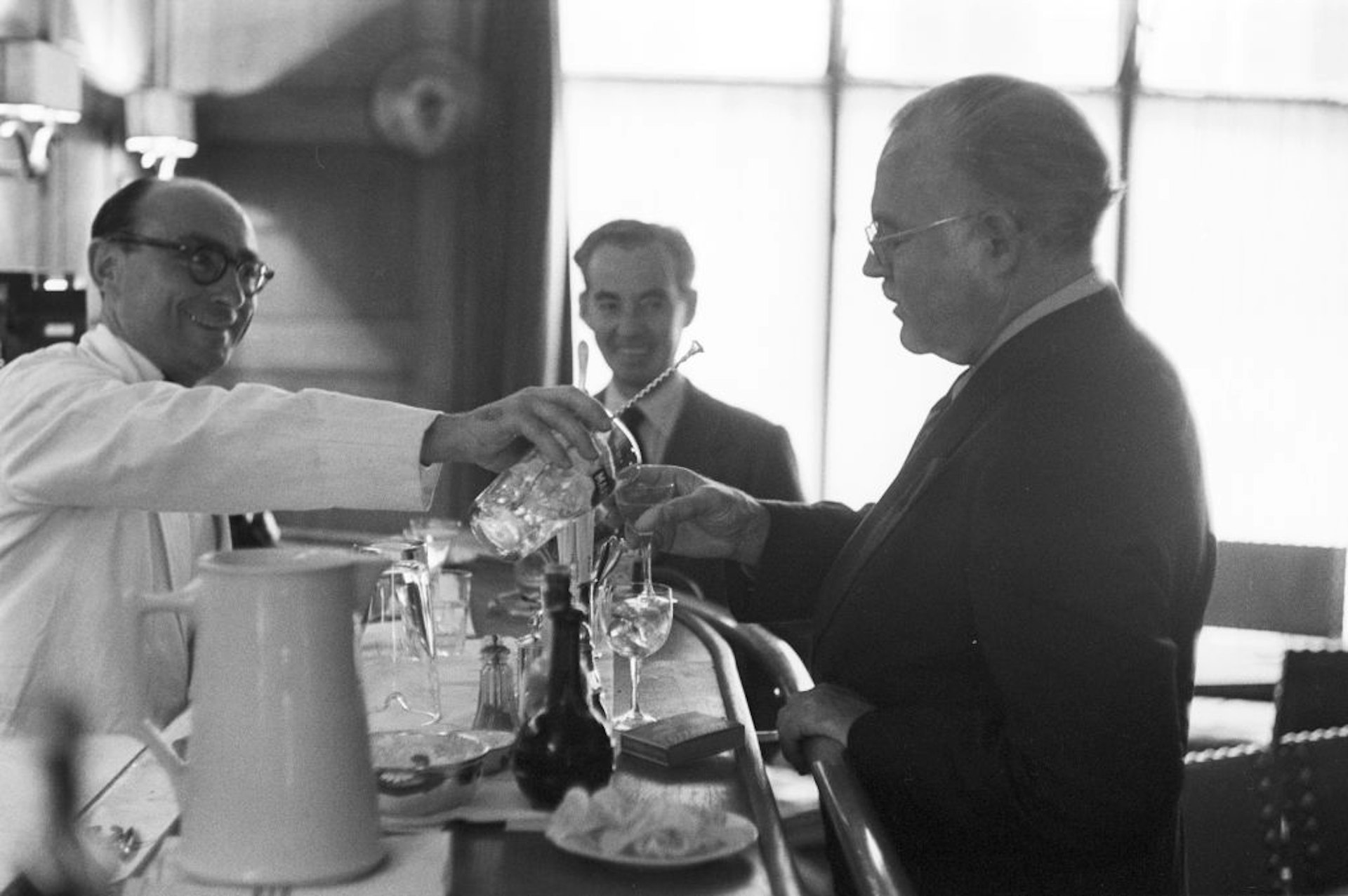 , Ernest HEMINGWAY aux courses hippiques d' Auteuil. Au bar, un serveur en veste blanche sert à boire à l' écrivain.