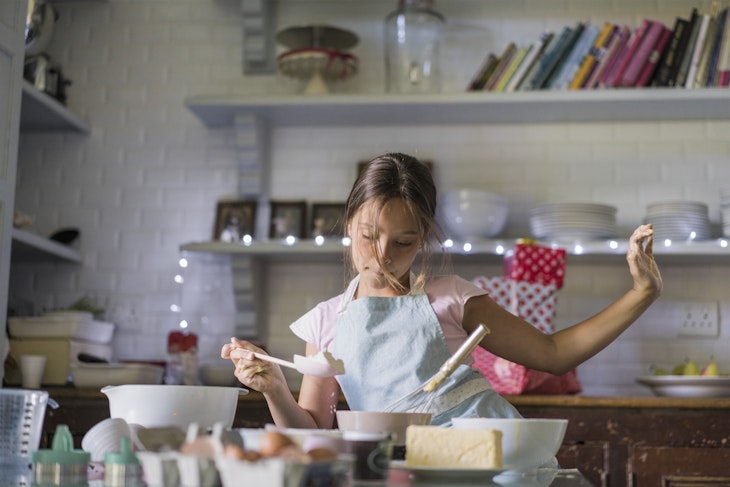 Girl is dancing while baking. Child is preparing food in kitchen. She is wearing apron.