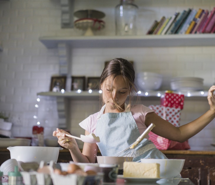 Girl is dancing while baking. Child is preparing food in kitchen. She is wearing apron.